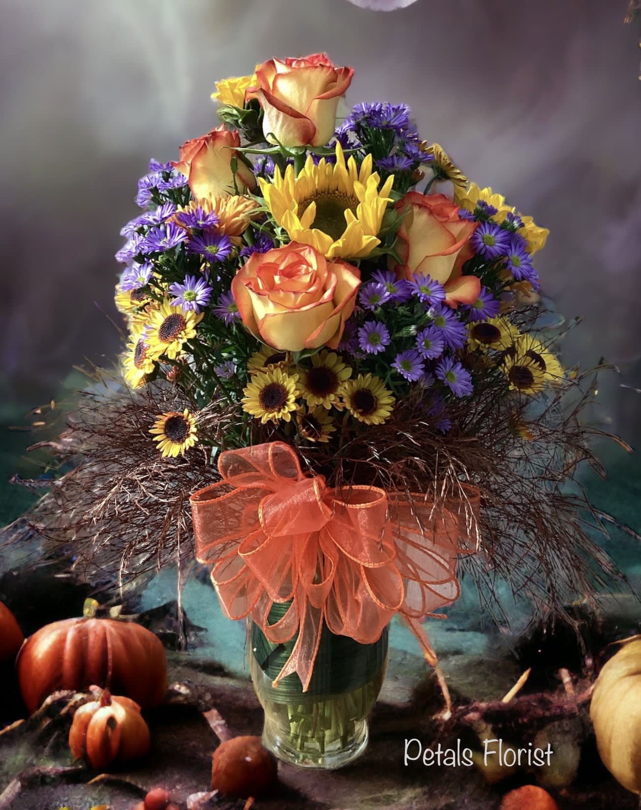 Vibrant Fall Bouquet by Petals  - Brighten any space with this stunning fall bouquet, a creative assortment of seasonal blooms selected to bring joy. The rich combination includes striking purple asters, fresh chrysanthemums, sunny sunflowers, and warm orange roses. Accented by deep burgundy tree fern and a sheer orange bow, it is elegantly displayed in a glass vase. We are confident you'll be pleased with this one-of-a-kind arrangement. Please call Cheryl with any questions at 401-828-0701. 