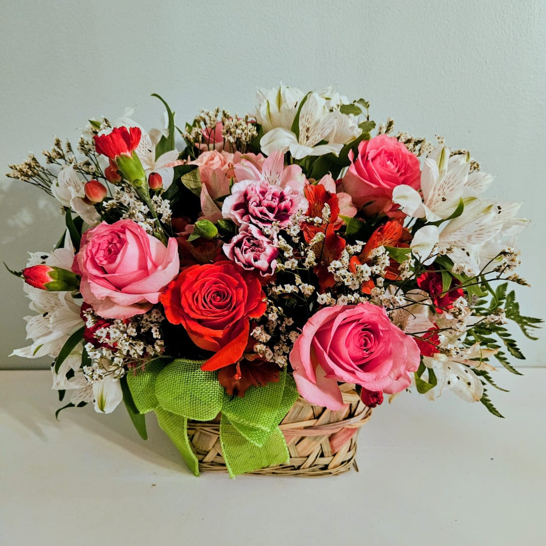 Mixed pink and red flower arrangement in a woven basket with a green ribbon