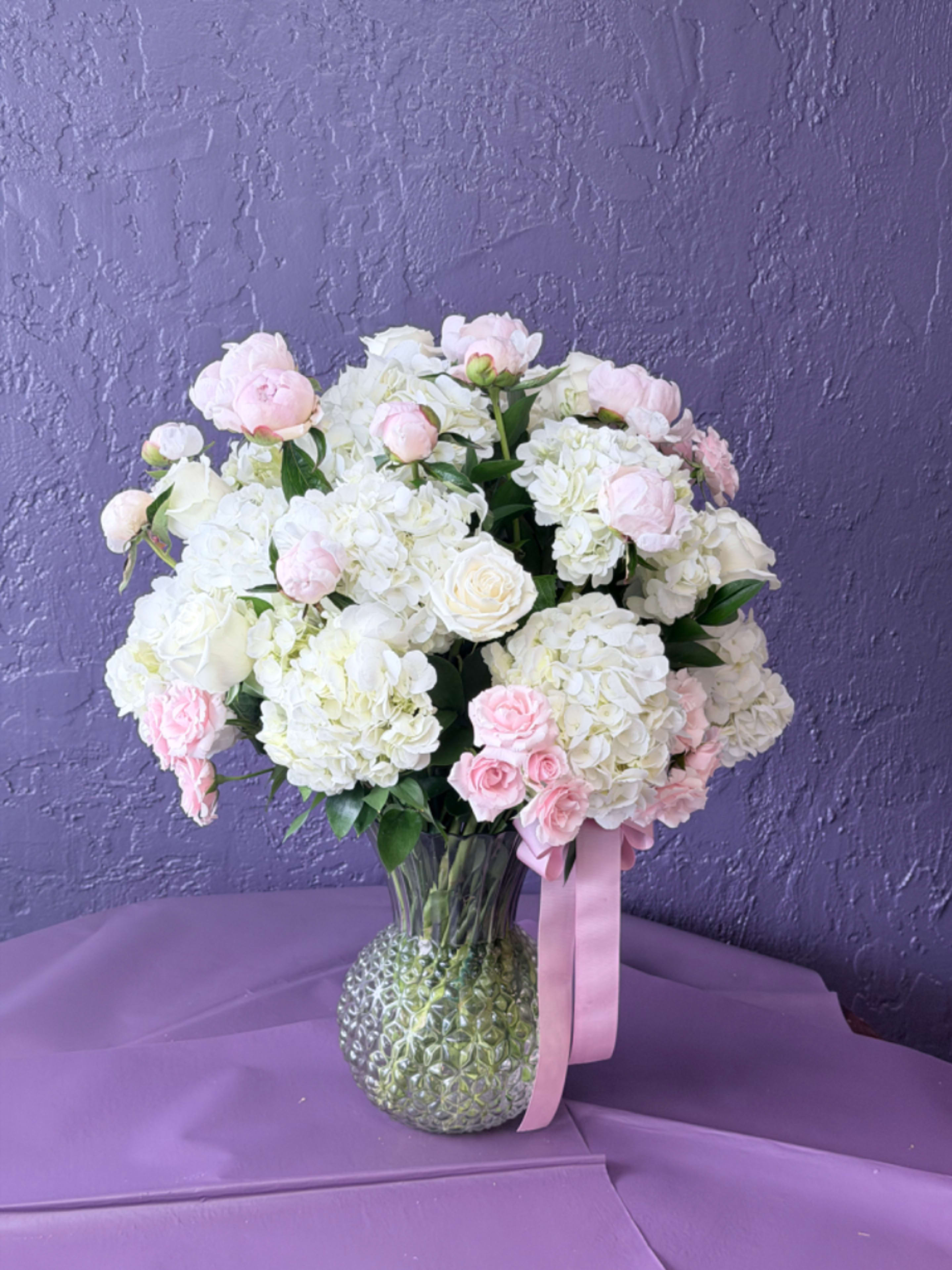 My Darling - A compact, garden-style bouquet that feels both romantic and cheerfully intimate, perfect as an anniversary or birthday flower arrangement to make her feel truly cherished. My Darling combines rounded roses and delicate ranunculus with peony-like blooms and airy chrysanthemums, so the person you're thinking of will notice the layered textures and soft, feminine palette right away-ideal for Mother's Day, Valentine's Day, or simply to say "congratulations" or "thinking of you."  Soft pinks and lavenders mingle with pops of red, orange, white hydrangea clusters, and fresh greenery, gathered tight in a clear glass vase and finished with a pale pink ribbon for a polished, petite presentation. The arrangement reads bright and elegant on a bedside table or small dining centerpiece; approximate dimensions 11" diameter x 11" height. Lightly recut stems and fresh water will keep the mix looking its best for several days-an effortless way to make her feel seen and celebrated.