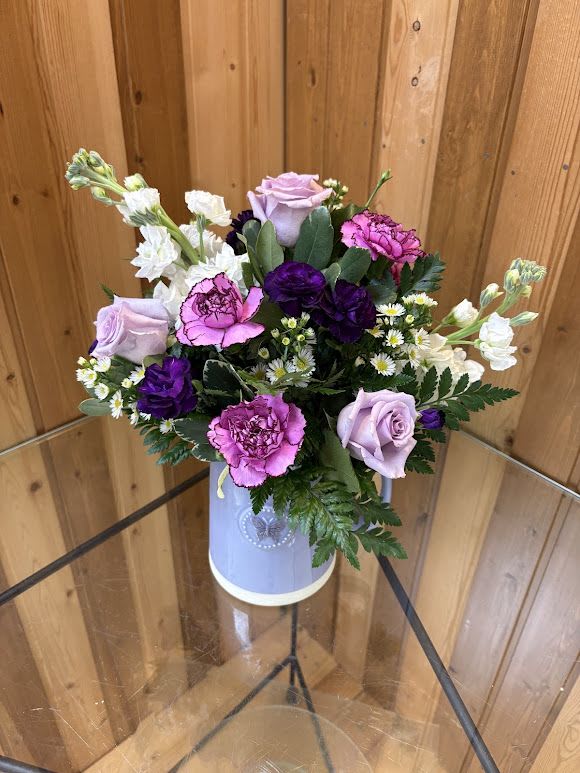 Lavender Garden Pitcher - This charming arrangement is designed in a beautiful purple ceramic pitcher, giving it a cozy garden-fresh feel that doubles as a keepsake container. Lavender roses, bright pink carnations, rich purple accent blooms, and delicate white daisies are gathered together with lush greenery to create a soft, romantic color palette.  The pitcher adds a touch of farmhouse charm while the mix of flowers brings texture, movement, and cheerful color. Once the flowers are enjoyed, the ceramic pitcher can be reused as décor, a vase, or even in the kitchen, making this gift both beautiful and lasting.  Lavender Pitcher Garden is perfect for birthdays, thank-you gifts, thinking-of-you moments, or simply brightening someone’s day with a fresh garden look. 