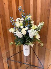 Serenity Cross Bouquet - A serene, upright sympathy arrangement that brings quiet comfort - the Serenity Cross Bouquet is designed for memorial services, funerals, and moments of faith. The tall white roses and trumpet-shaped lilies form a gentle, reverent centerpiece while soft blue delphinium and pale yellow stock add calm accents that lift the composition. Presented in a smooth white ceramic vase embossed with a simple cross, this bouquet reads as a thoughtful expression of sympathy and spiritual solace for the person you're thinking of.  The design features rounded white blooms clustered through the center with taller, slender blue delphinium rising above for vertical movement and an airy silhouette. Pale yellow stock threads through the arrangement to introduce warmth and subtle contrast against the white and blue palette. Vase dimensions and exact flower counts are available at checkout; if any flower is unavailable we may substitute with blooms of equal or greater value and similar color-always chosen to preserve the arrangement's tranquil look.  Ideal for a graveside service, church memorial, or to send to a family at home, this bouquet conveys reverence and gentle support so the recipient feels comforted and seen. Haehn Florist, Greenhouses, &amp; Flower Delivery - 410 Hamilton Rd. Wapakoneta. Follow us at www.facebook.com/haehnflorist for updates and seasonal offerings.
