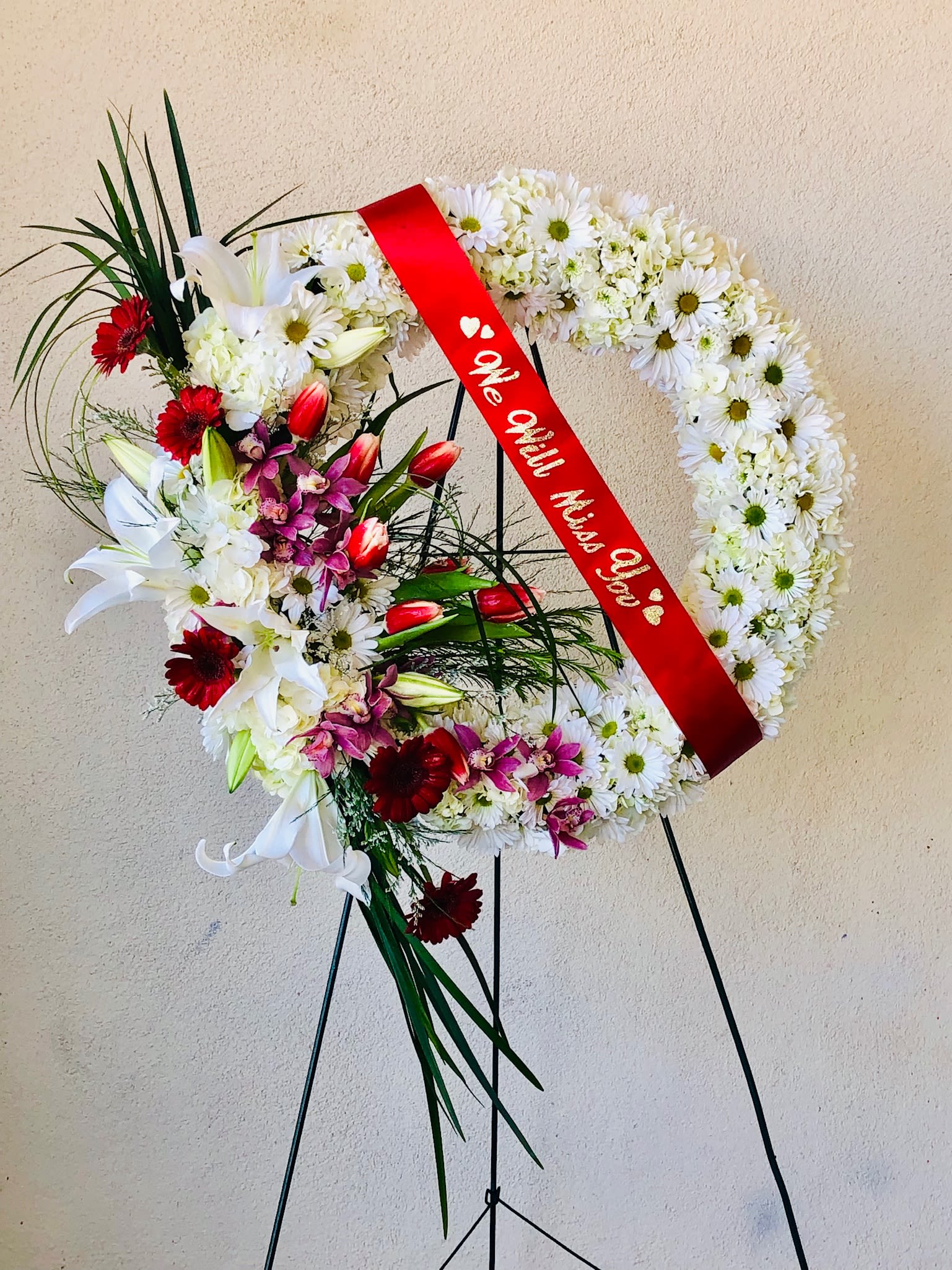 Heart-shaped funeral wreath with white daisies, lilies, and red ribbon on a stand