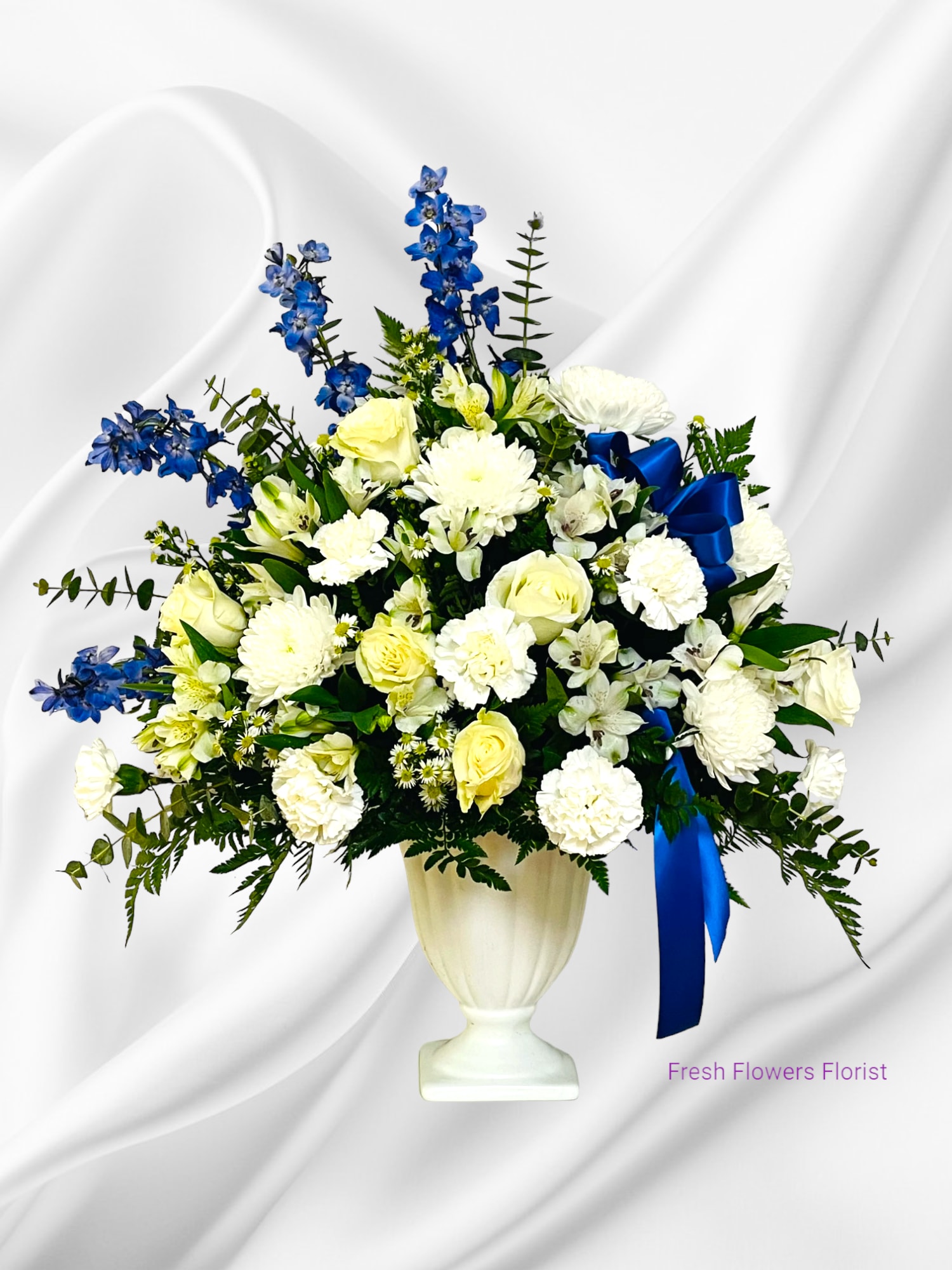 White and yellow floral arrangement with blue flowers in a white urn vase
