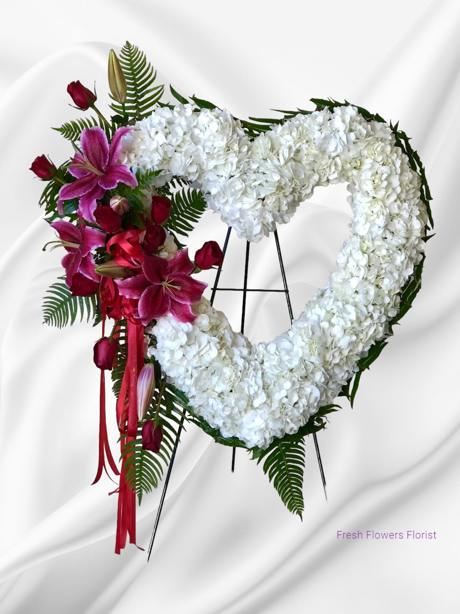 Heart-shaped white floral wreath with pink lilies and red roses on an easel