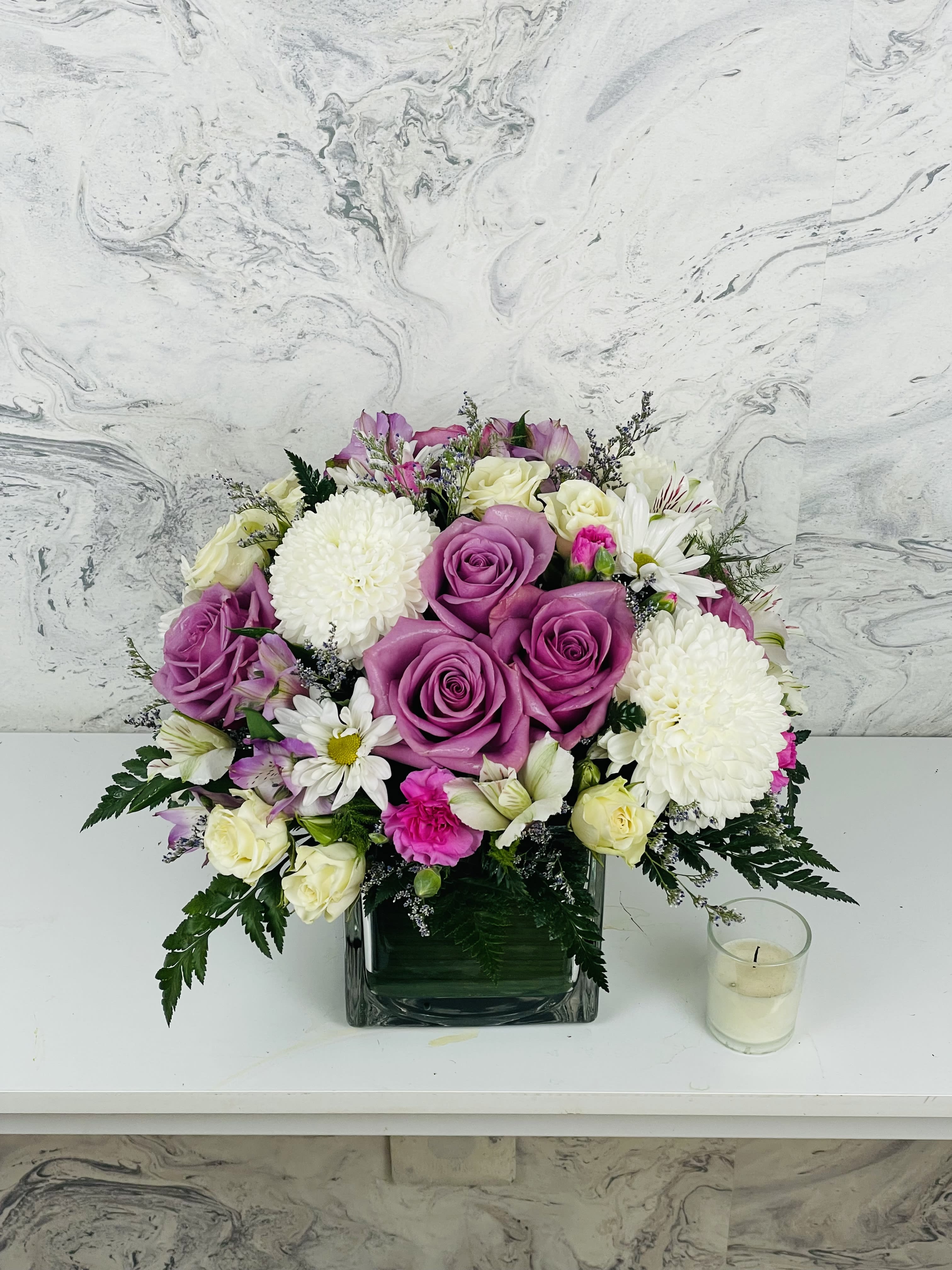 Pink and white rose bouquet in a square glass vase with a small candle beside it.