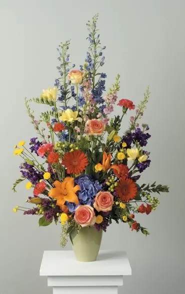 Tall mixed flower arrangement in a pale vase with orange, pink, yellow, and blue blooms