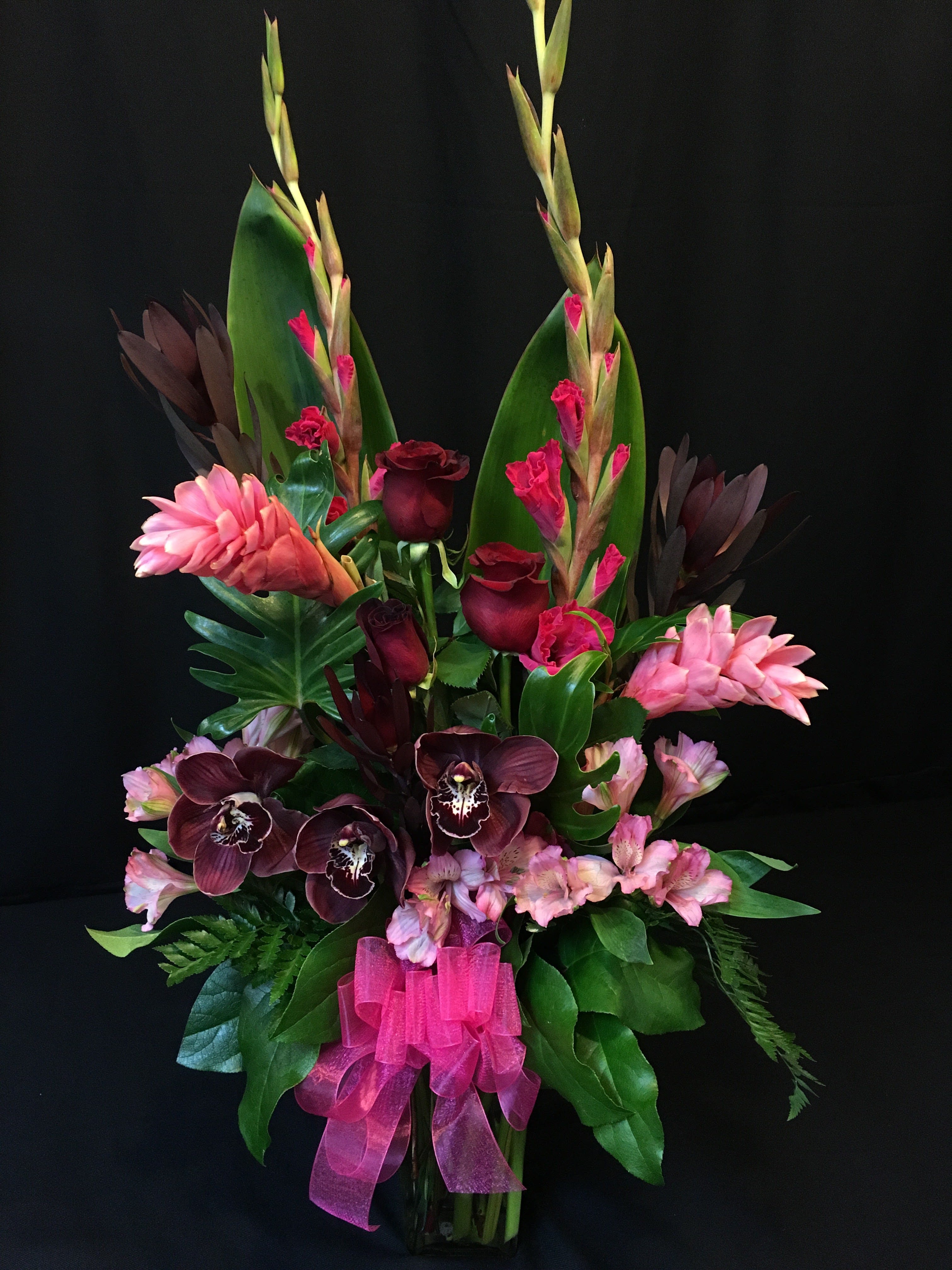 Tropical bouquet with pink orchids, dark roses, and a pink ribbon in a vase