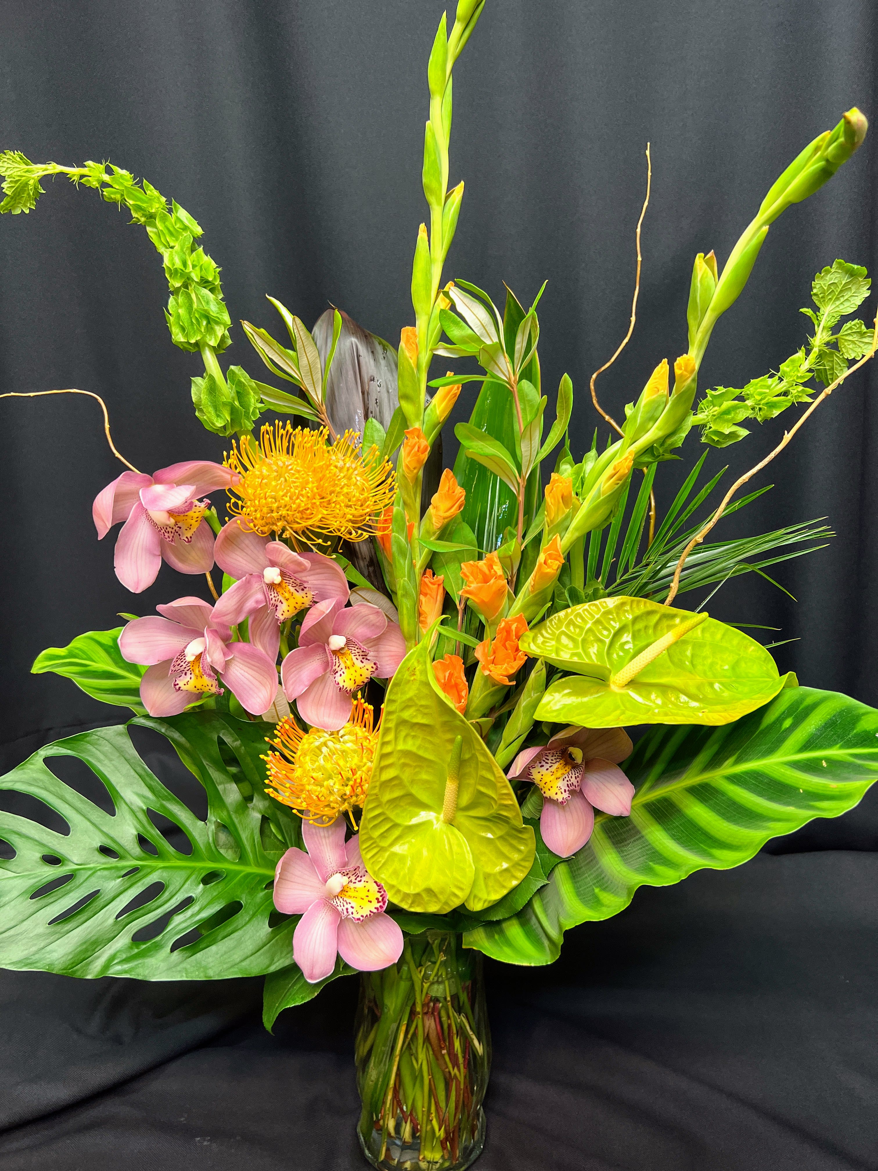 Tropical arrangement with pink orchids, yellow pincushion blooms, and anthuriums in a glass vase