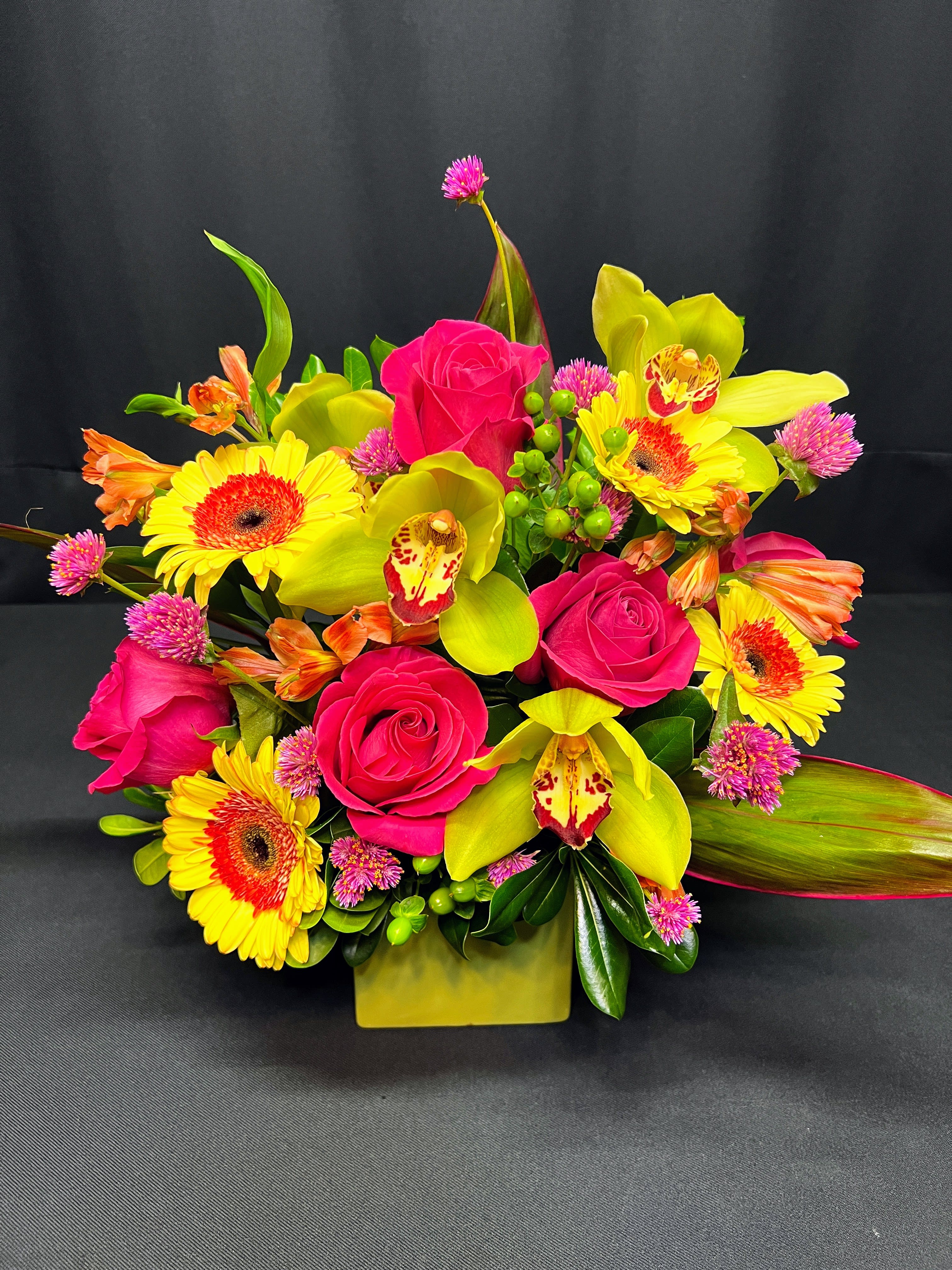 Bright bouquet of pink roses, yellow orchids, and gerbera daisies in a square vase