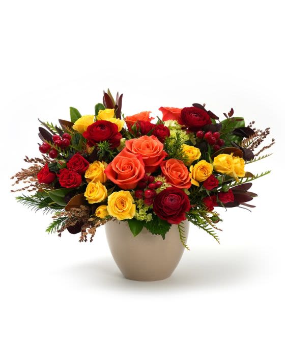 Low arrangement of red, orange, and yellow roses with ranunculus in a beige ceramic vase.