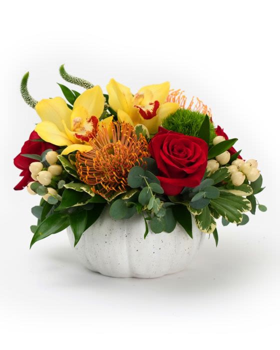 Low arrangement of yellow orchids, orange protea, and red roses in a white pumpkin-shaped bowl