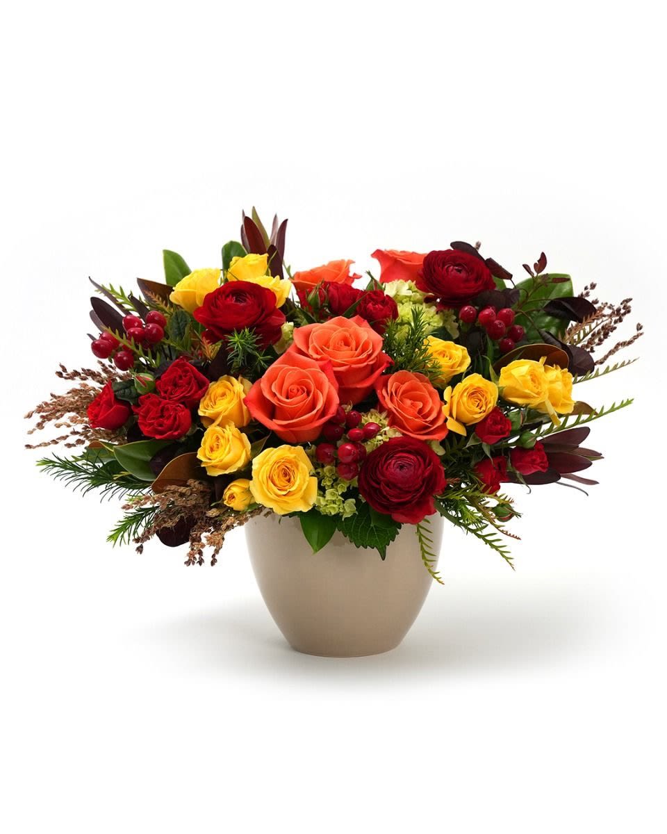 Low arrangement of orange, red, and yellow roses in a beige ceramic vase with red berries