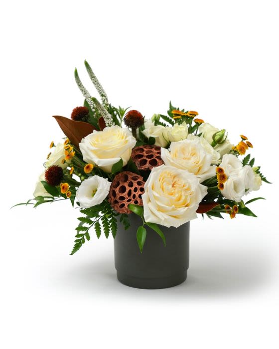 Low arrangement of cream roses, white blooms, and yellow accents in a dark cylinder vase with lotus pods.