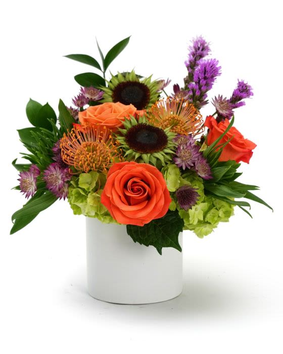 Compact arrangement of orange roses, green hydrangeas, and purple blooms in a white cylinder vase