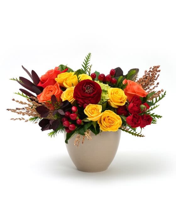 Low arrangement of yellow, orange, and red roses with berries in a beige vase