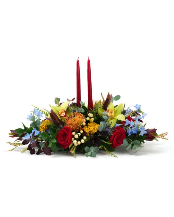 Low centerpiece with red roses, yellow pincushion blooms, green orchids and blue flowers around two tall red candles