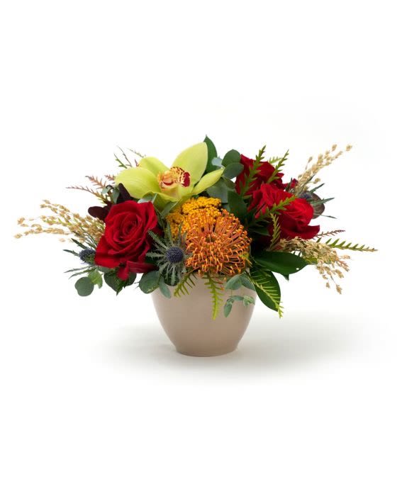 Low arrangement of red roses, green cymbidium orchid, and orange protea in a beige bowl vase