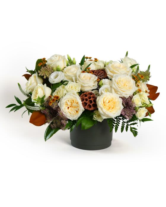 Low arrangement of cream roses with brown seed pods in a dark cylinder vase