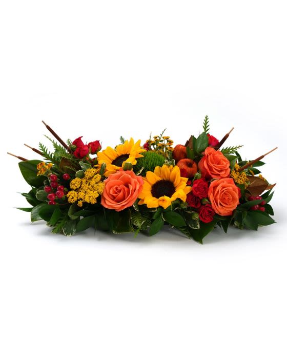 Low centerpiece of orange roses, yellow sunflowers, and red accent blooms in an elongated design