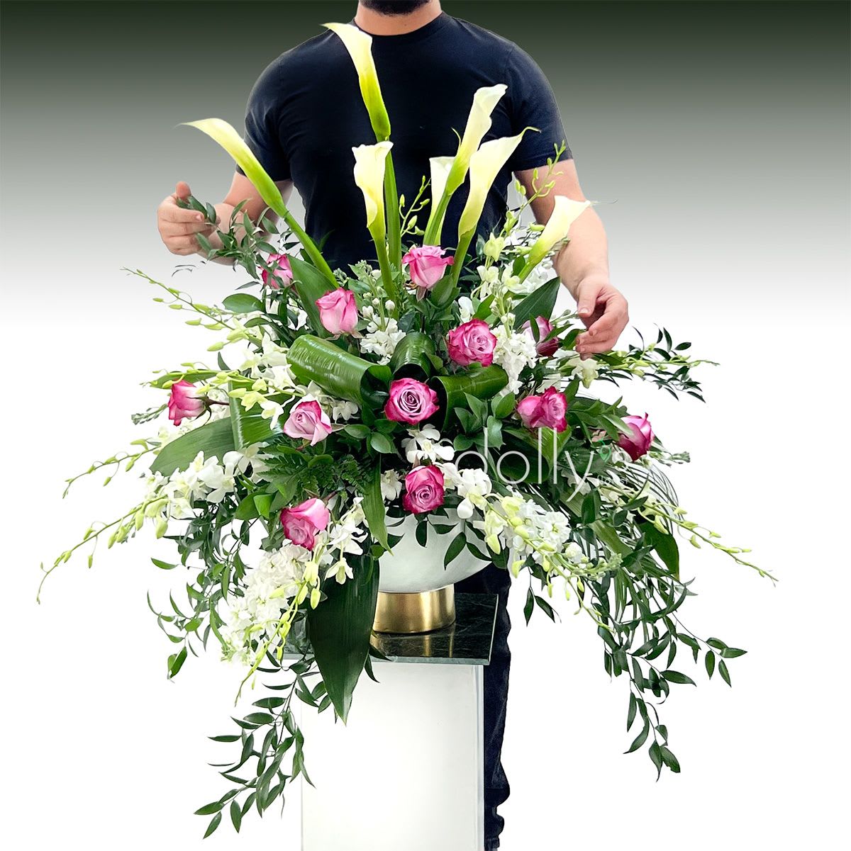 Empress - Towering white calla lilies rise above a lush cloud of magenta roses and white orchids in this large, dramatic flower arrangement-perfect as a refined birthday bouquet, elegant thank-you gift, or an extraordinary centerpiece for major celebrations. The contrast of magenta and crisp white against deep green foliage creates a polished, luxurious impression that will make the person you're sending it to feel truly celebrated.  Designed to spread broadly with cascading greenery and arching stems, the arrangement sits in a white ceramic vase with gold gilding. The mix features calla lilies, magenta roses, orchids, complimented with green leaves, delivers a formal, sculptural silhouette that reads as everyday luxury. Ideal for someone who appreciates classic elegance and dramatic height- ready to display and makes an impressive arrival.
