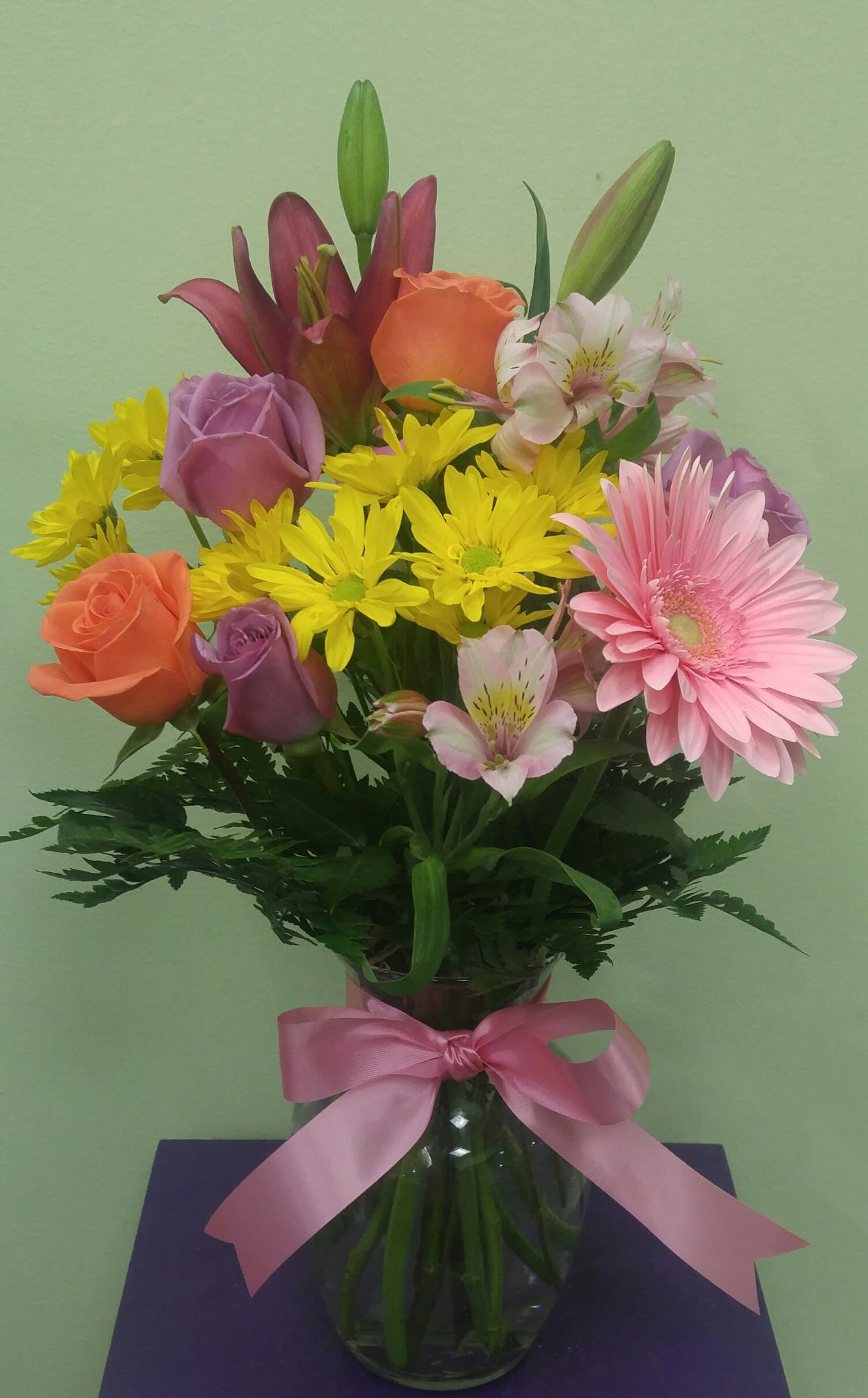 Mixed bouquet of roses, lilies, daisies, and alstroemeria in a glass vase with a pink ribbon