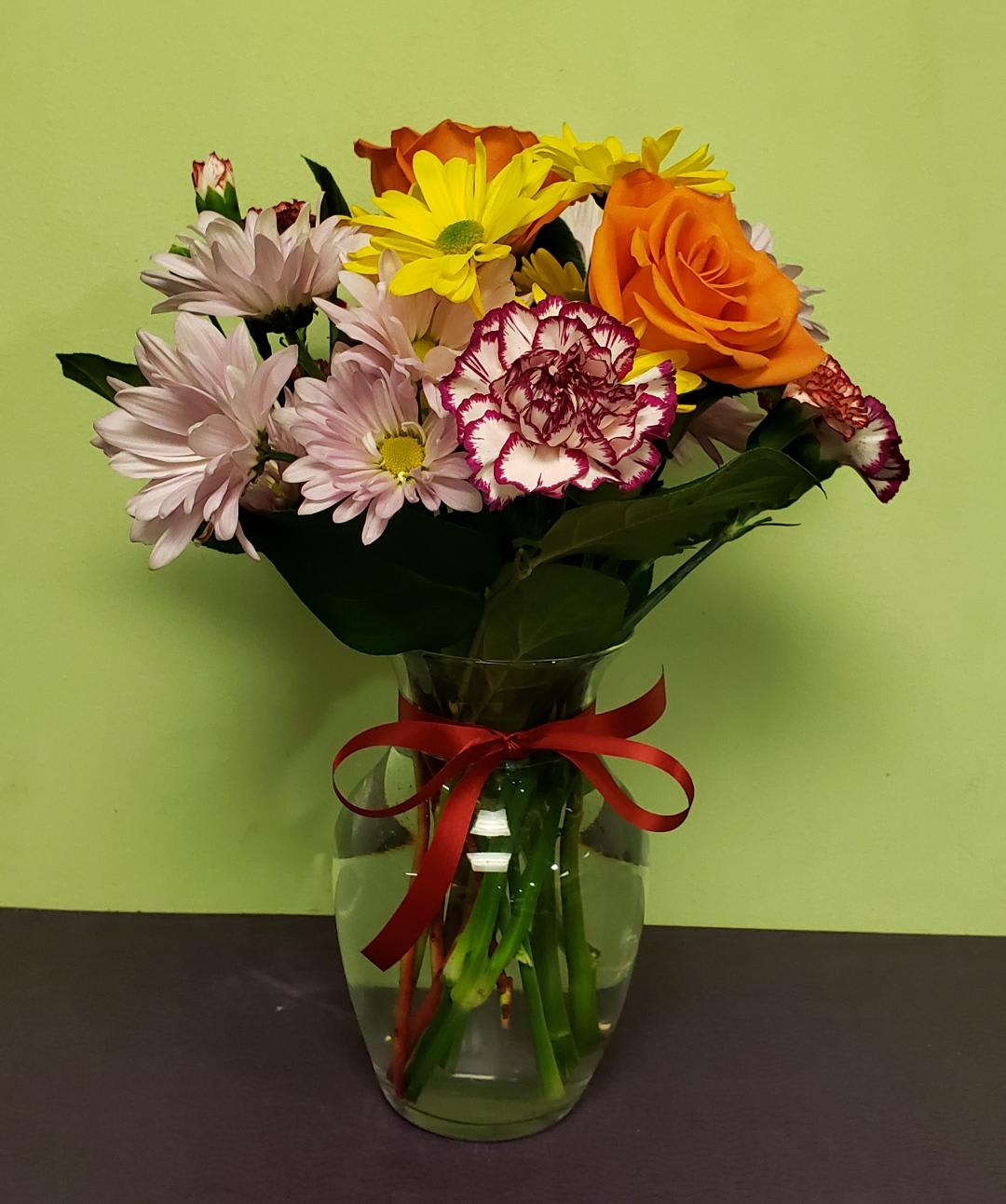 Mixed bouquet of daisies, roses, and carnations in a glass vase with a red ribbon