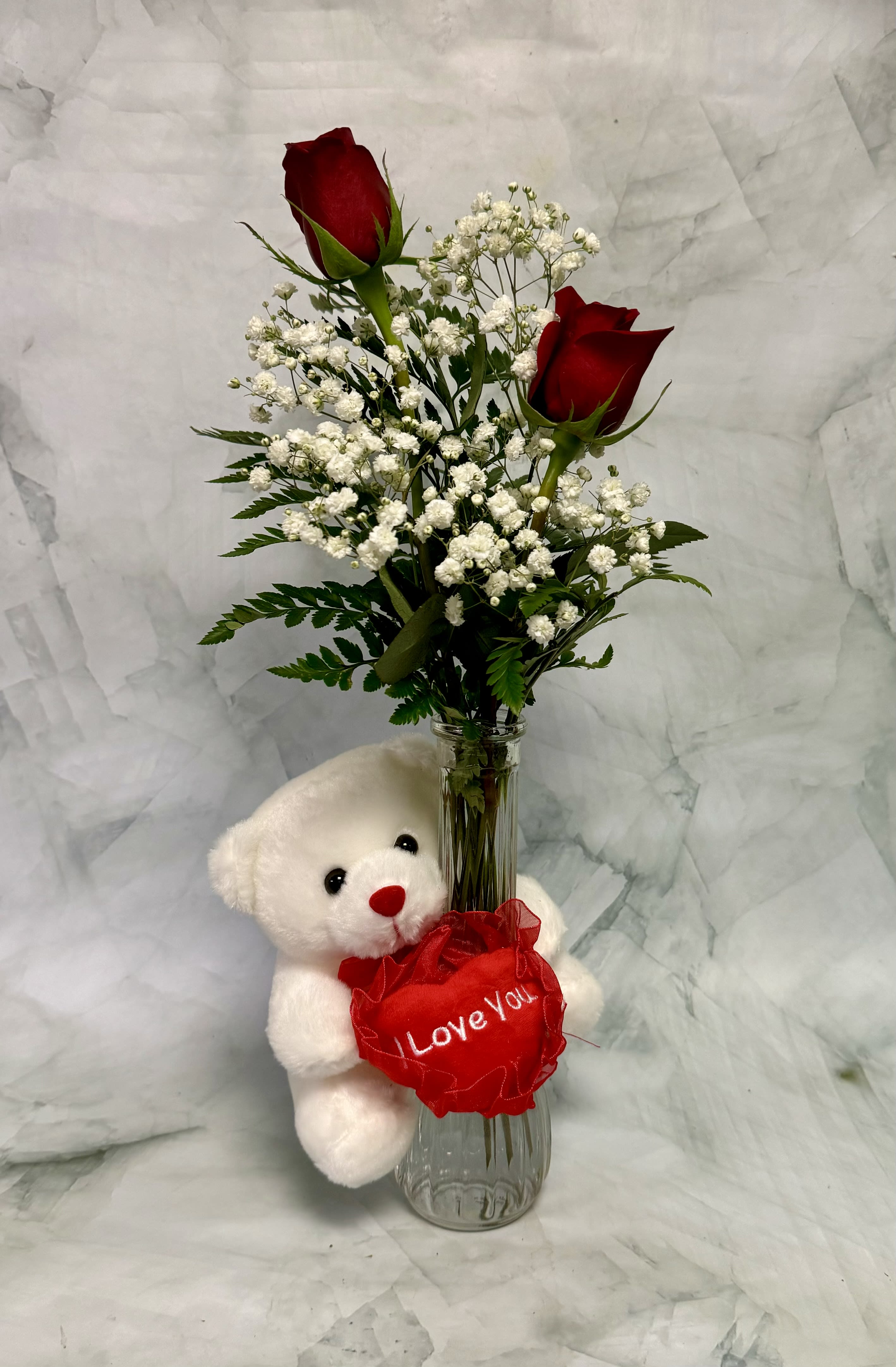 Two red roses in a glass vase with a teddy bear holding a heart