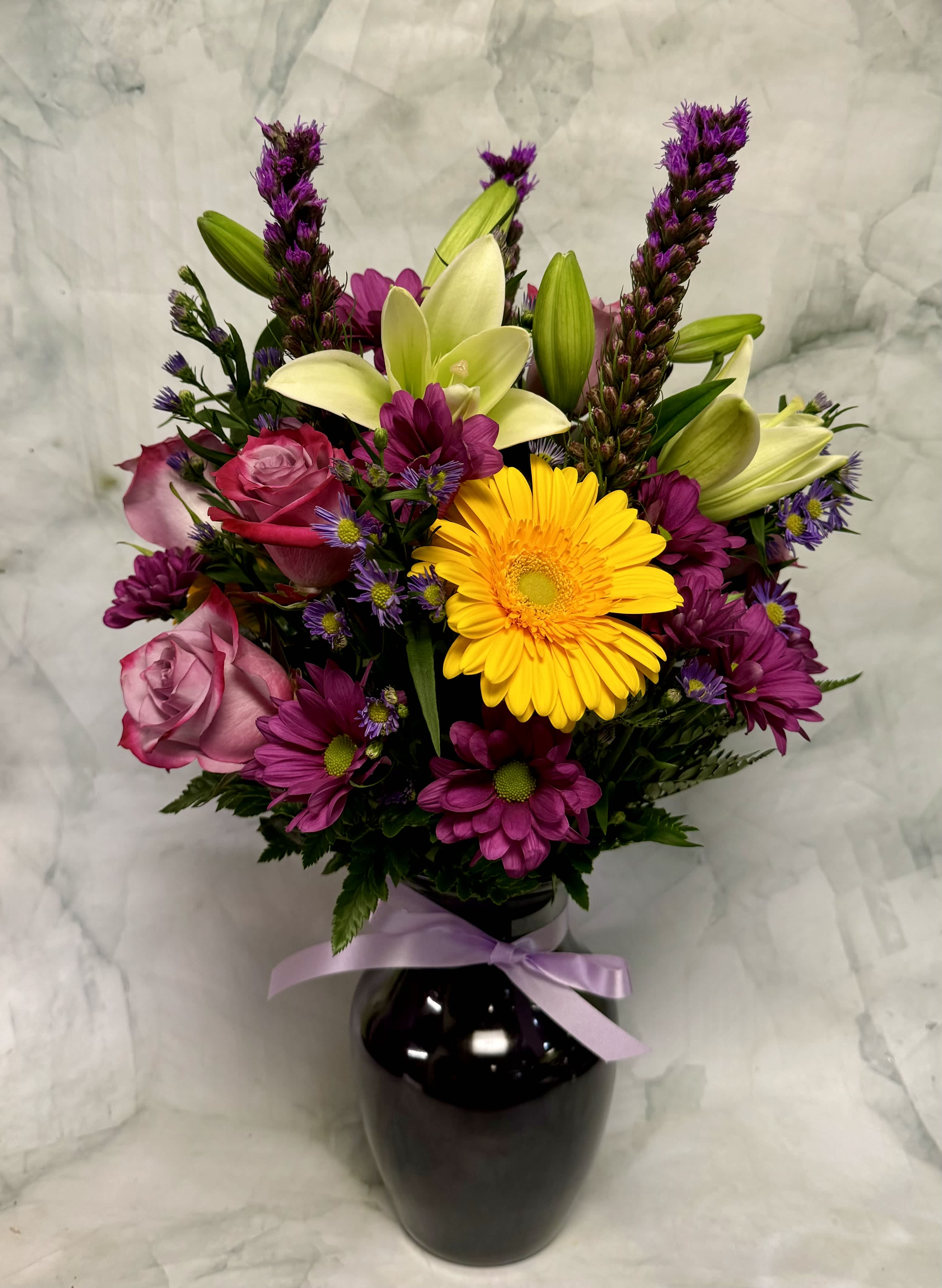 Mixed bouquet of roses, lilies, and daisies in a black vase with a lavender ribbon