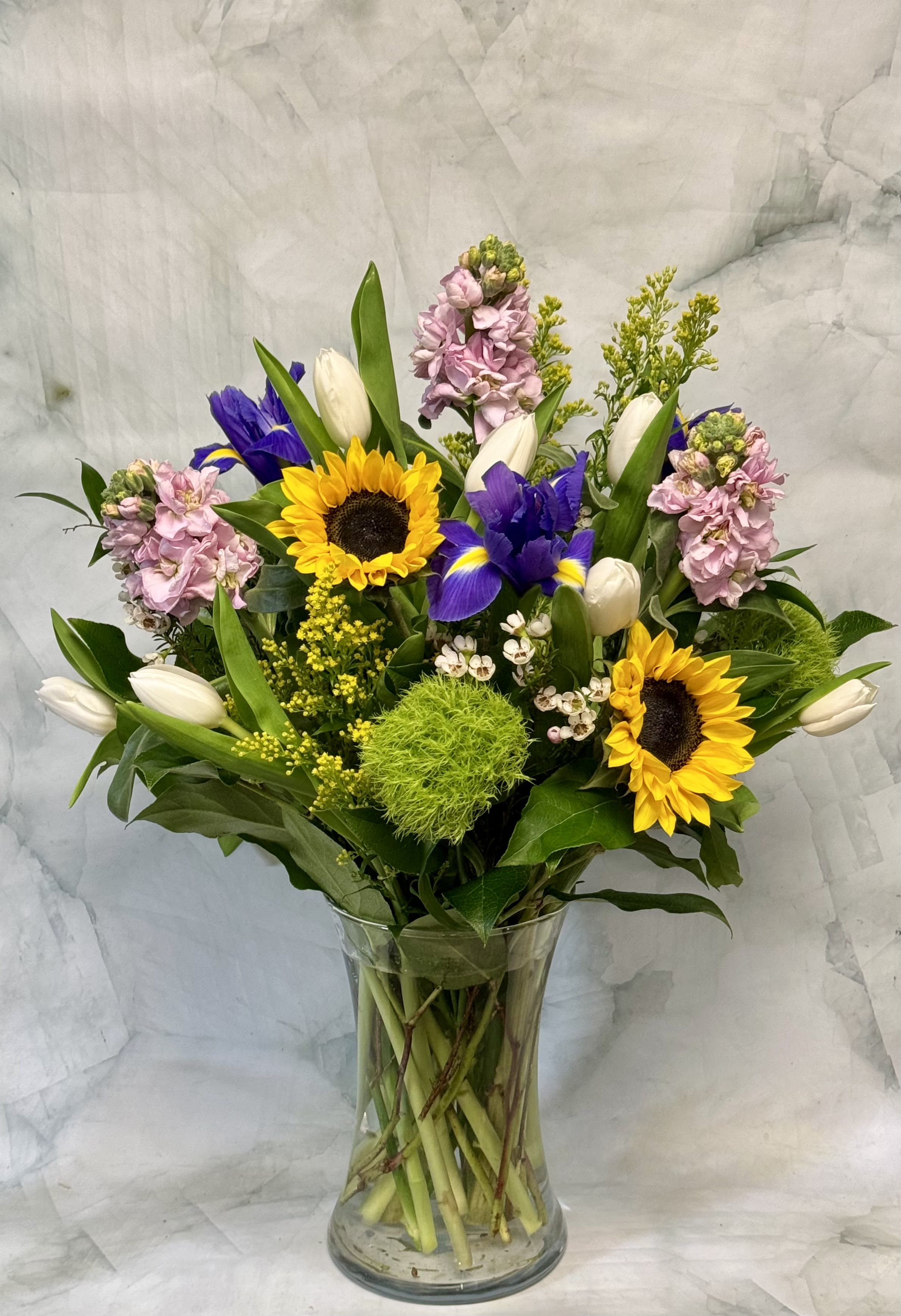 Mixed bouquet with sunflowers, purple irises, white tulips, and pink blooms in a glass vase