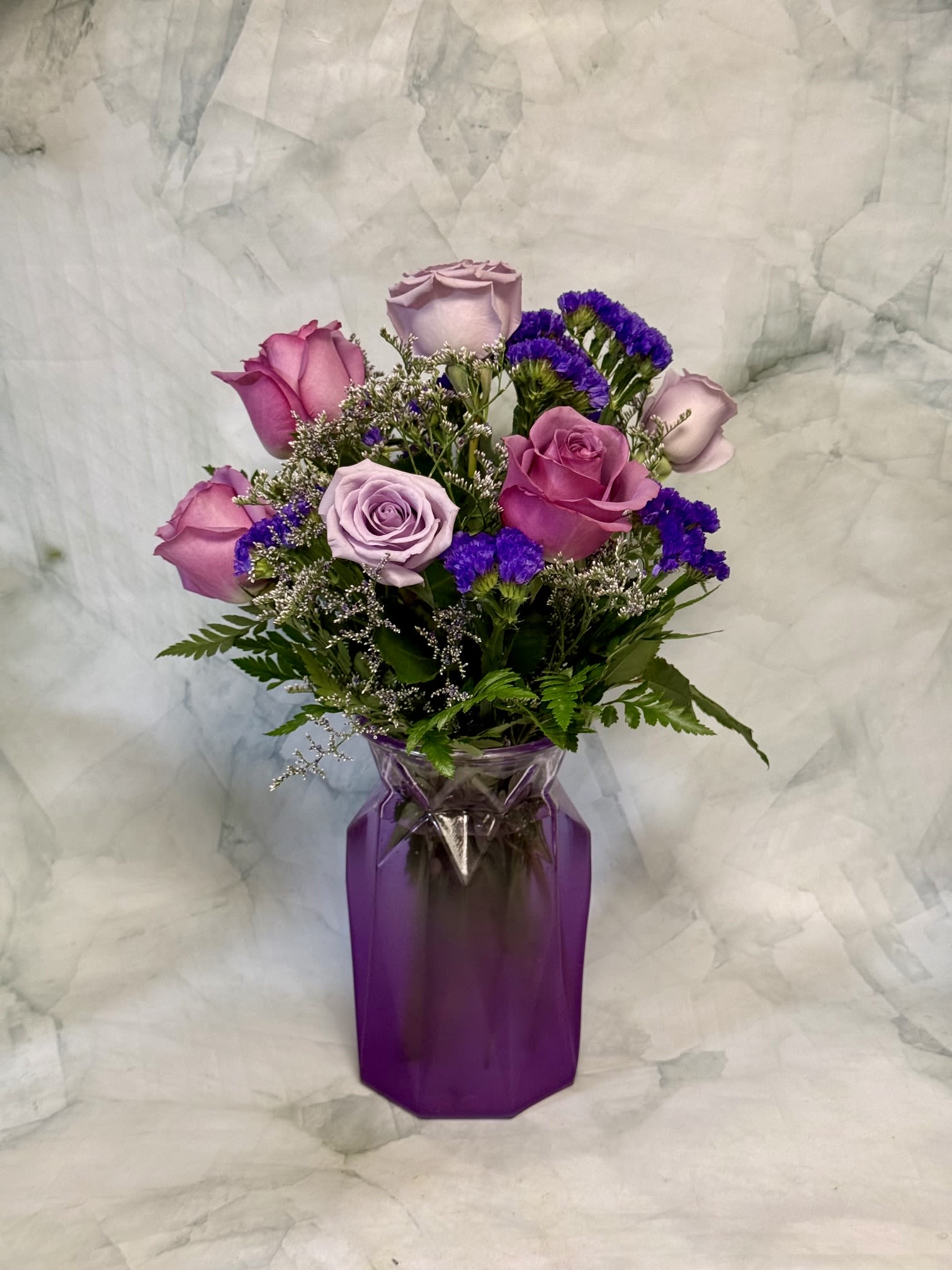 Lavender Love  - Soft lavender and pink roses mingle with delicate purple fillers and airy white accents to create a romantic birthday bouquet or anniversary arrangement that feels made for Mom. This flower arrangement for Mother's Day, a birthday, or "just because" moments presents a gentle, sentimental palette-pink, purple, white, and green-that reads both classic and modern, so the person you're thinking of will feel cherished and seen.  The blooms are gathered in a tall purple glass vase that gives the design a rich, contemporary base and makes the softer rose tones pop. Texturally, the smooth rose petals contrast with the feathery white accents and small purple filler flowers for a polished, layered look. Sized to sit as a focal gift on a table or mantel, it arrives assembled and ready to display; simple fresh-water care-change water every couple days and trim stems if needed-keeps the roses looking their best.