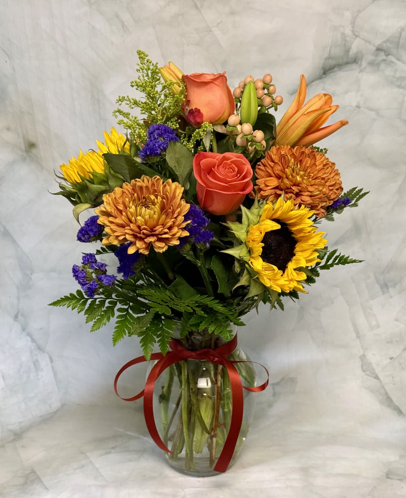 Mixed bouquet of sunflowers, roses, and orange blooms in a glass vase with a red ribbon