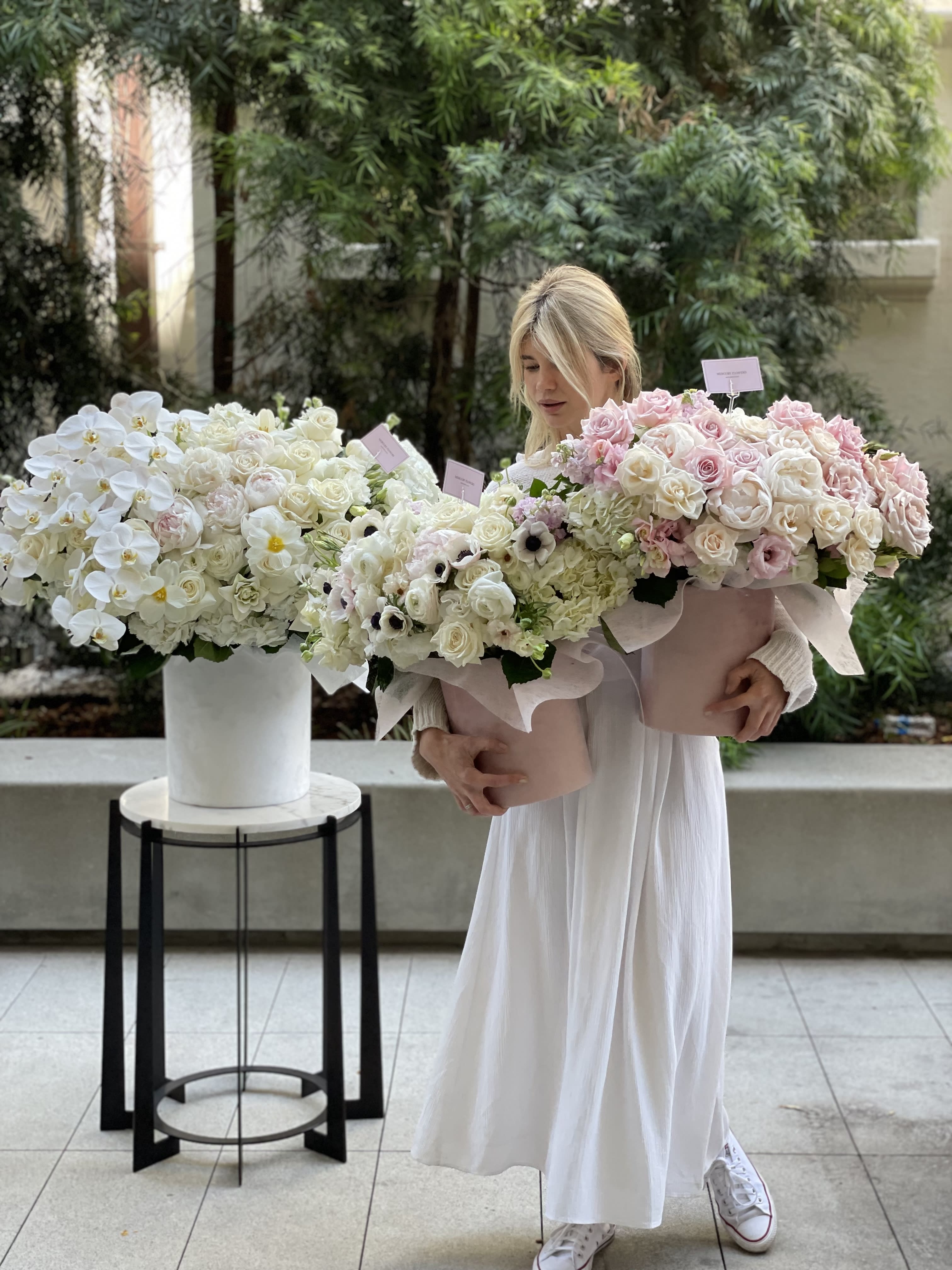 Woman holding two large pastel flower bouquets beside a white floral arrangement.