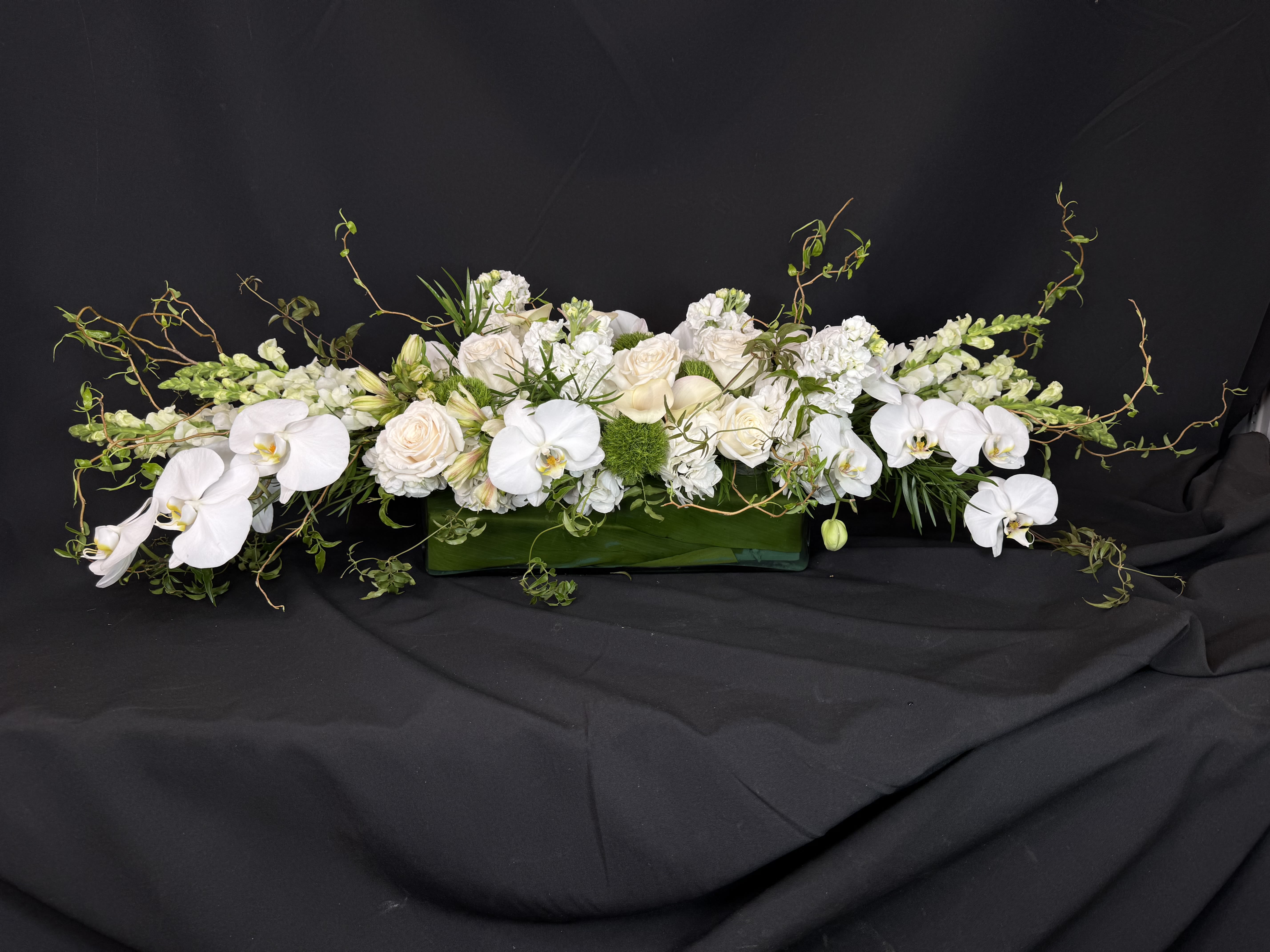 Long white floral arrangement with orchids and roses in a green rectangular container