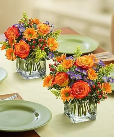 Two orange and purple floral centerpieces in square glass vases on a table