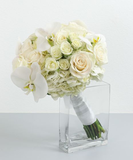 White bridal bouquet of roses, hydrangea, and orchids with ribbon-wrapped stems in a clear glass vase