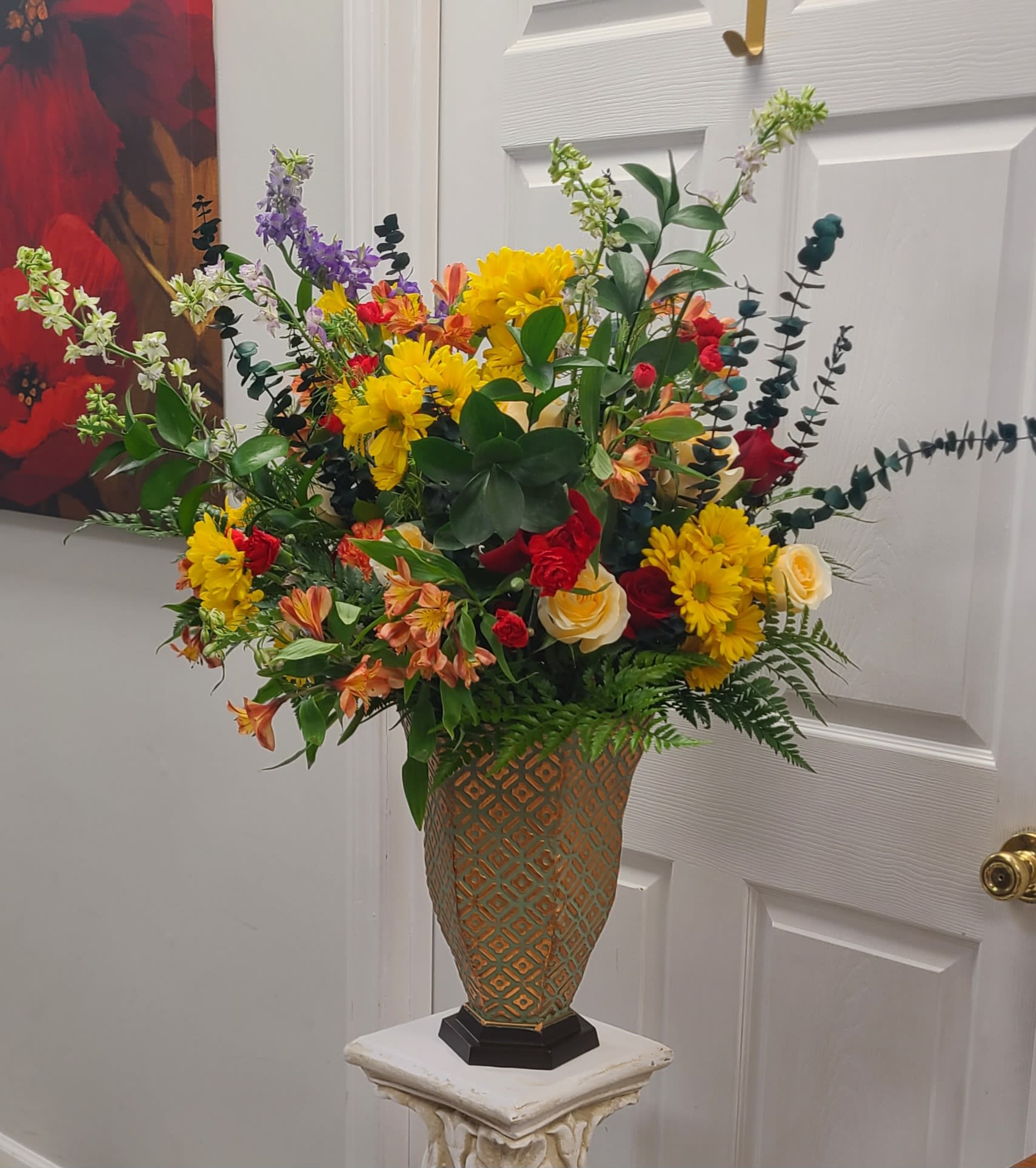 Event Ready  - A towering, celebratory mixed flower arrangement built to catch the eye - perfect as a birthday bouquet, congratulations gift, or uplifting centerpiece for a special event. Bright yellow chrysanthemums and daisy-like blooms set a sunny, energetic tone, while red roses and orange alstroemeria bring warmth and passion; tall purple and white spike flowers add dramatic height and movement so the person you're thinking of will feel truly celebrated the moment it arrives.  This large, upright design is densely layered with glossy foliage, branching stems, cream roses, snapdragon-like spikes and stock for texture, all arranged in a patterned decorative vase set on a pedestal stand. The palette of yellow, red, orange and purple reads festive and full-bodied, with a garden-style, airy silhouette that fills a room. Ideal for birthdays, congratulations, thank-you gifts, or to brighten a home, it arrives ready to display - no fussing required.