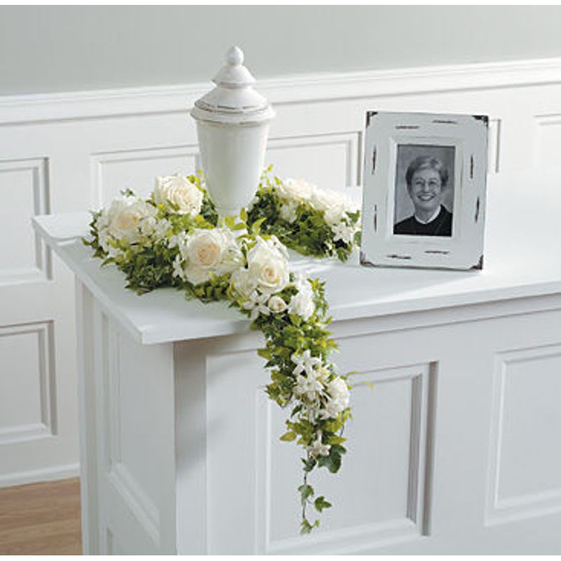 White funeral urn surrounded by white roses and greenery beside a framed portrait