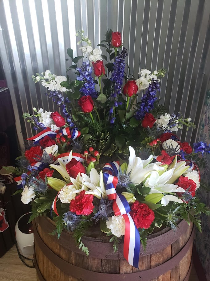 Eternal Patriot Tribute - This is a Eternal Patriotic Tribute for Cremation Urn Arrangement, designed to honor and celebrate the life of a loved one, particularly someone with a connection to military service, public service, or a strong sense of national pride. Description: Color Scheme: Red, white, and blue — representing patriotism and the national flag. Flowers Used: Red Roses &amp; Carnations – symbolize love, admiration, and remembrance. White Lilies &amp; Carnations – traditionally represent purity, peace, and spiritual rebirth. Blue Delphinium &amp; Thistle – bring depth and calm, often used to express dignity and grace. White Stock – symbolize a happy life and lasting affection. Hypericum Berries – add texture and natural richness to the arrangement. Special Features: Tri-Color Ribbon (Red, White, and Blue) – artistically tied throughout the arrangement, reinforcing the patriotic theme. Urn Ring Design – arranged in a circular fashion to surround a cremation urn (not present in the image), creating a focal point for the memorial.