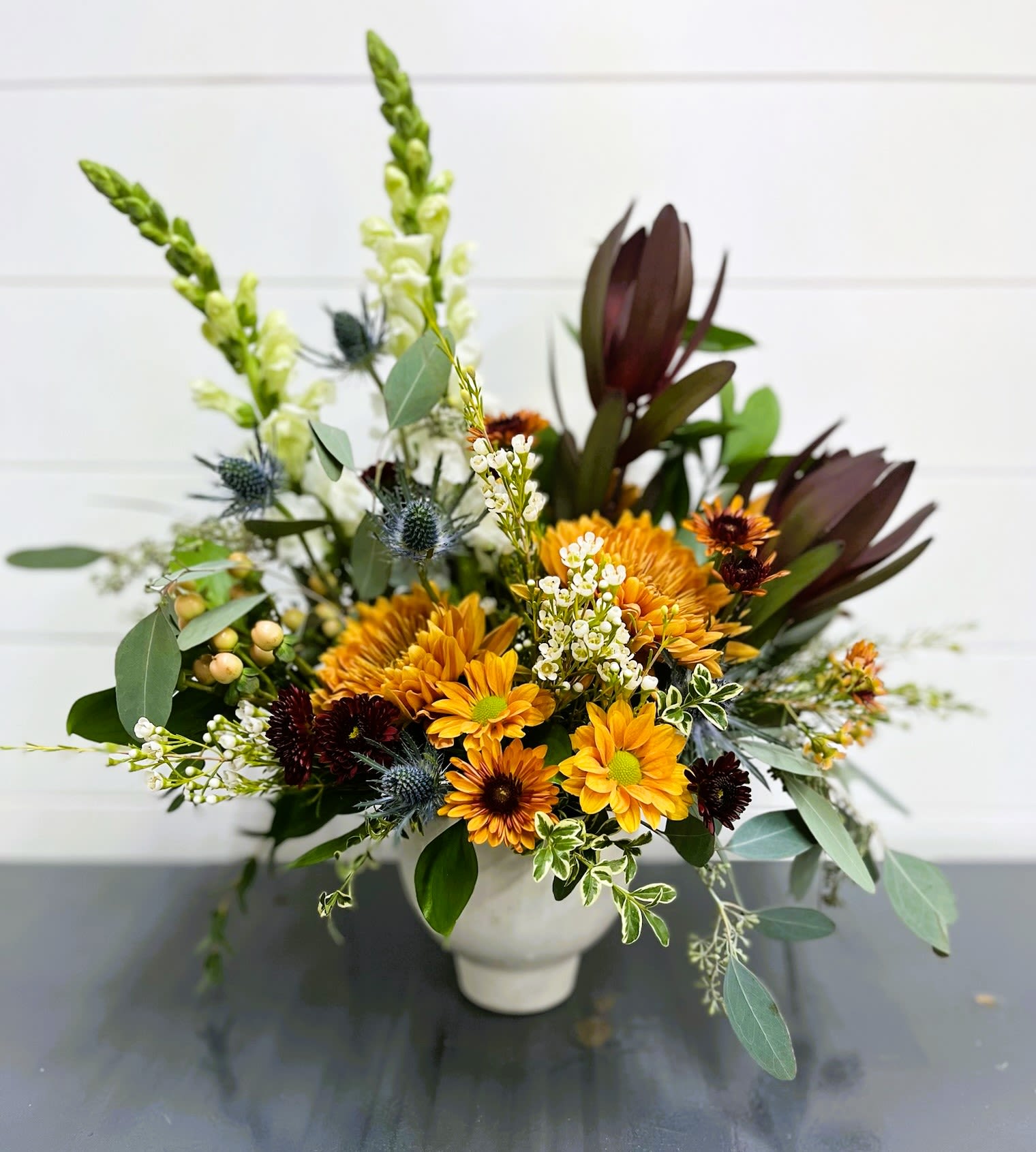 Modern Autumn Sunset - This modern Autumn arrangement would look beautiful in any location in your home. The white ceramic container contains seasonal mums, Safari Sunset, peach hypericum berries, white waxflower, blue thistle, white stock and seasonal greenery. 