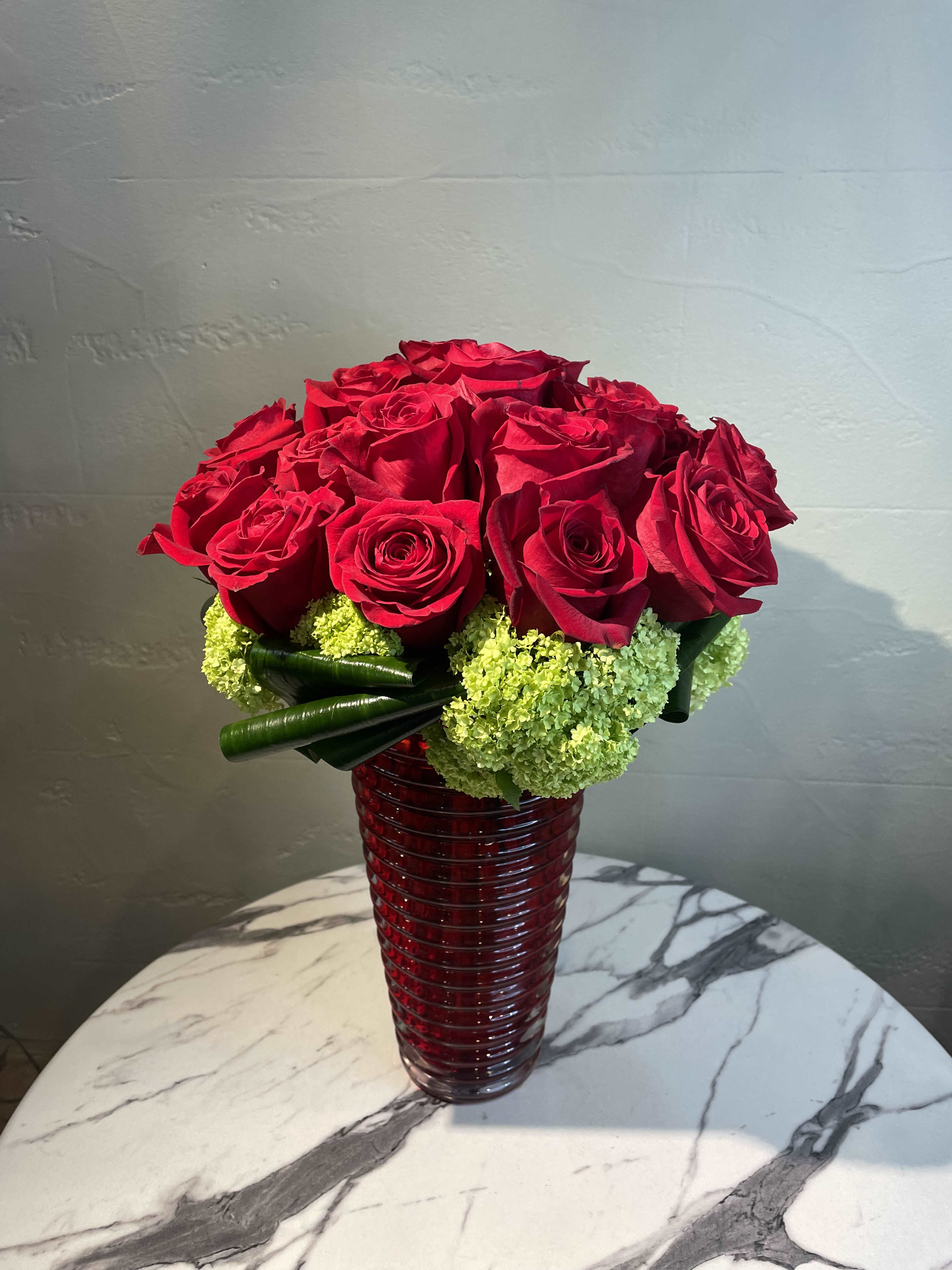 Red roses arranged in a tall red glass vase with green accents