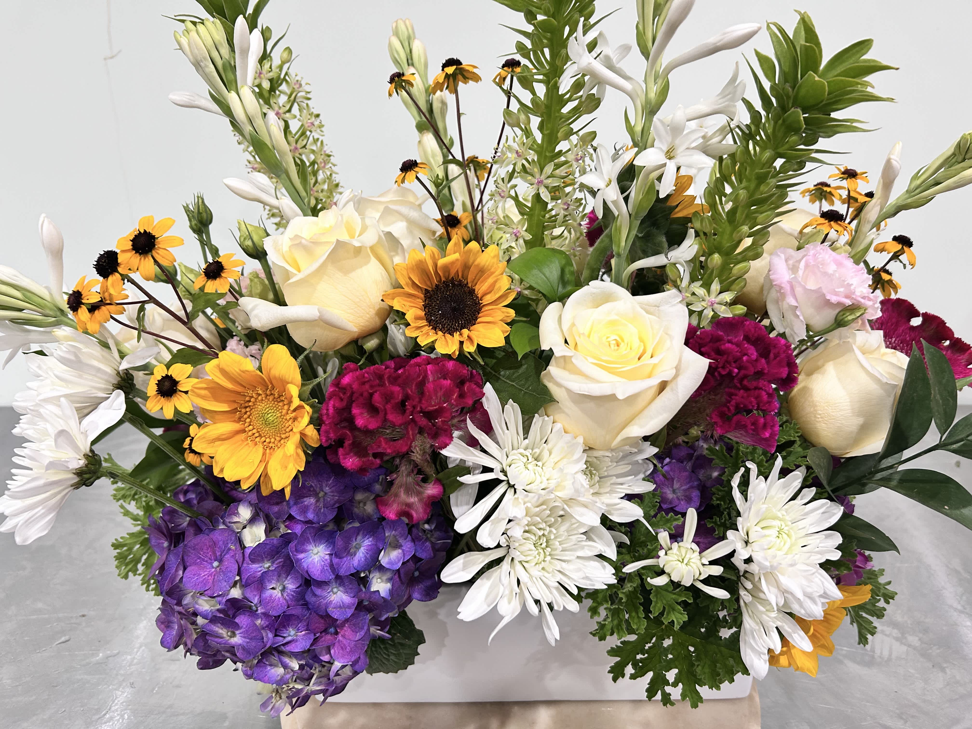 Mixed bouquet with white roses, purple hydrangeas, and yellow daisy-like flowers in a white container.