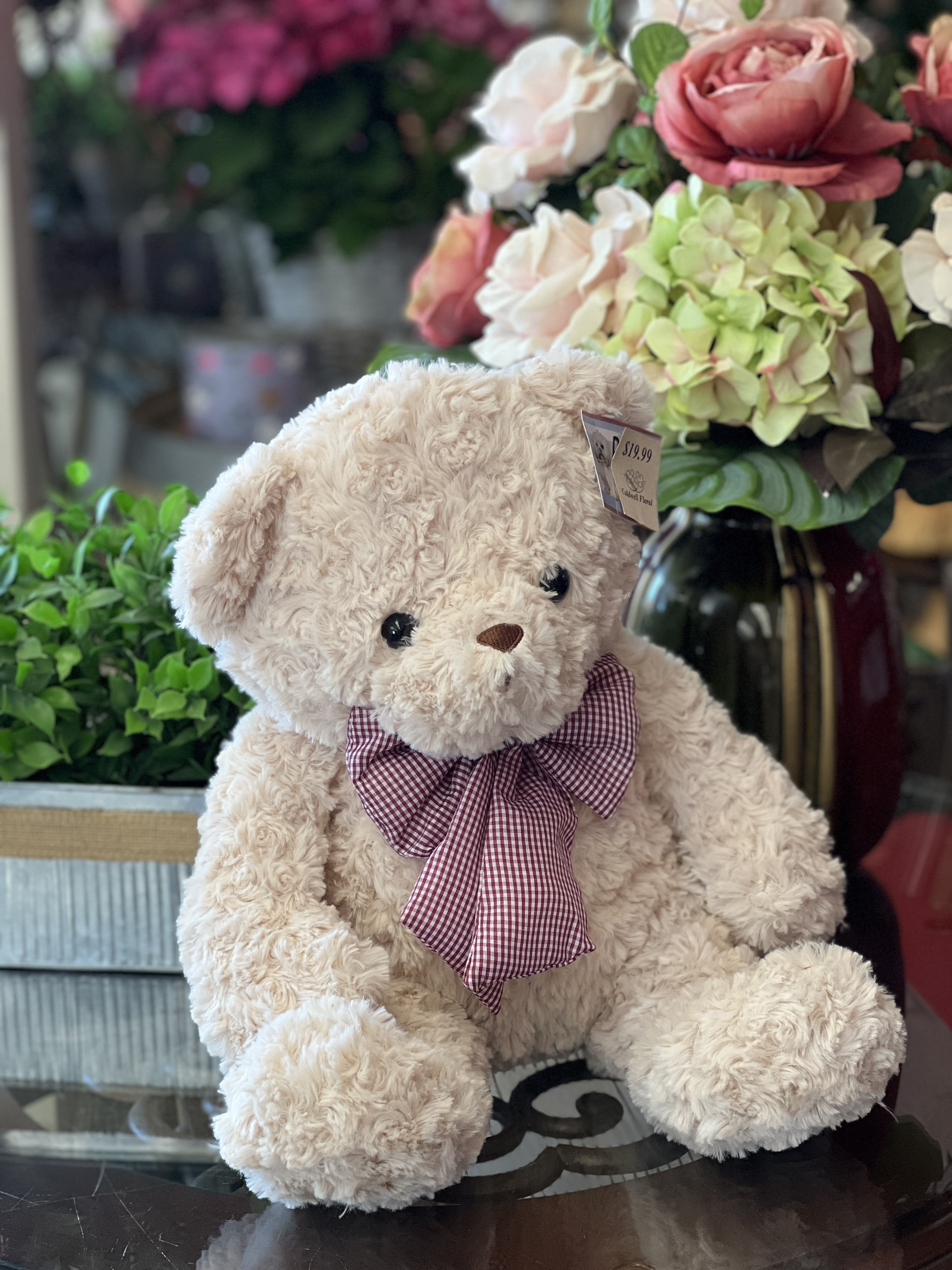 Plush teddy bear with a plaid bow sitting beside a floral arrangement