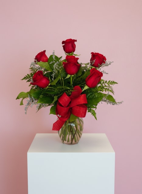 Red rose arrangement in a clear glass vase with a red bow on a white pedestal and pink background