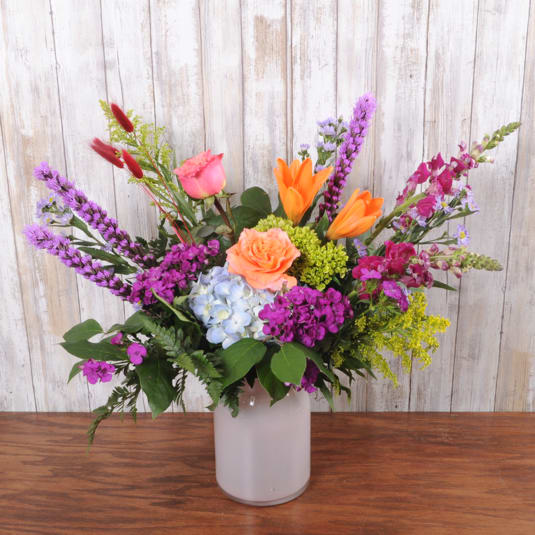 Garden Blend Pink - A lively, garden-style flower arrangement bursting with pink and peach roses, orange lilies, a pale blue hydrangea, and tall purple accents-perfect as a birthday bouquet, cheerful thank-you, or everyday gift that makes someone feel truly celebrated. The loose, asymmetrical design sends stems reaching outward from a simple white ceramic vase for a fresh, spontaneous look that reads as joyful and welcoming.  This full arrangement blends warm oranges and bright purples with soft blue and pink tones for a balanced, energetic palette. Roses and lilies provide fragrance and focal blooms, the hydrangea adds soft volume, and the taller purple flowers give height and movement. It arrives ready to display in the included white ceramic vase; for best enjoyment, trim stems and change the water every few days to keep blooms looking their freshest.