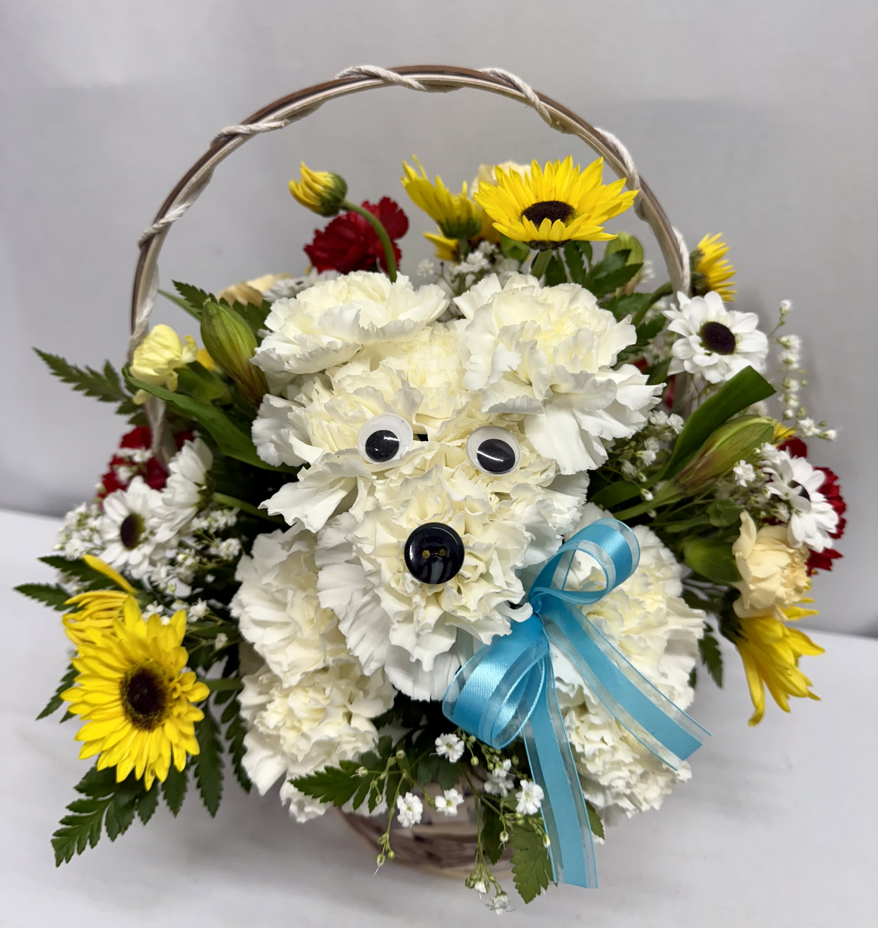 Basket arrangement with white carnation dog, yellow sunflowers, red and white daisies and a blue bow