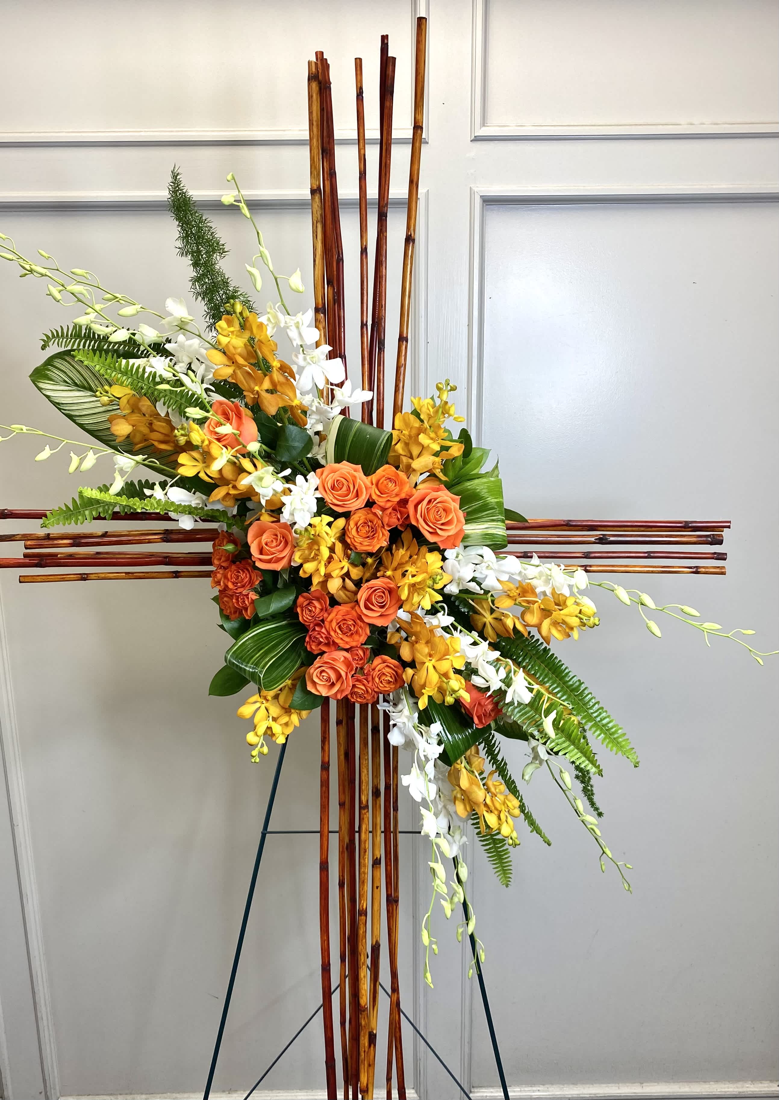 Tall cross-shaped bamboo standing spray with orange roses, yellow and white orchids, and tropical foliage.