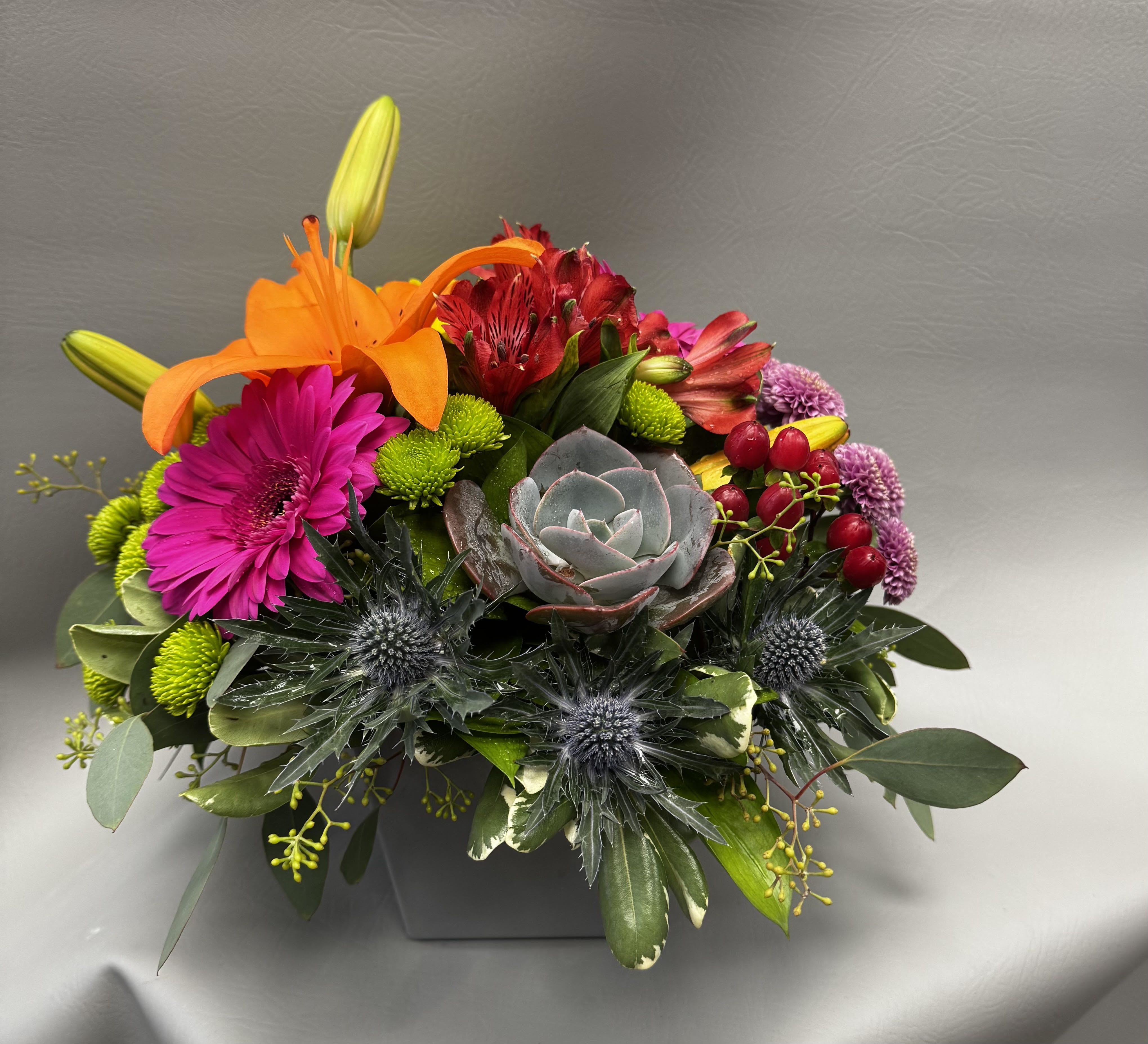 Colorful low arrangement with orange lilies, pink gerbera, succulents and mixed blooms in a gray square vase