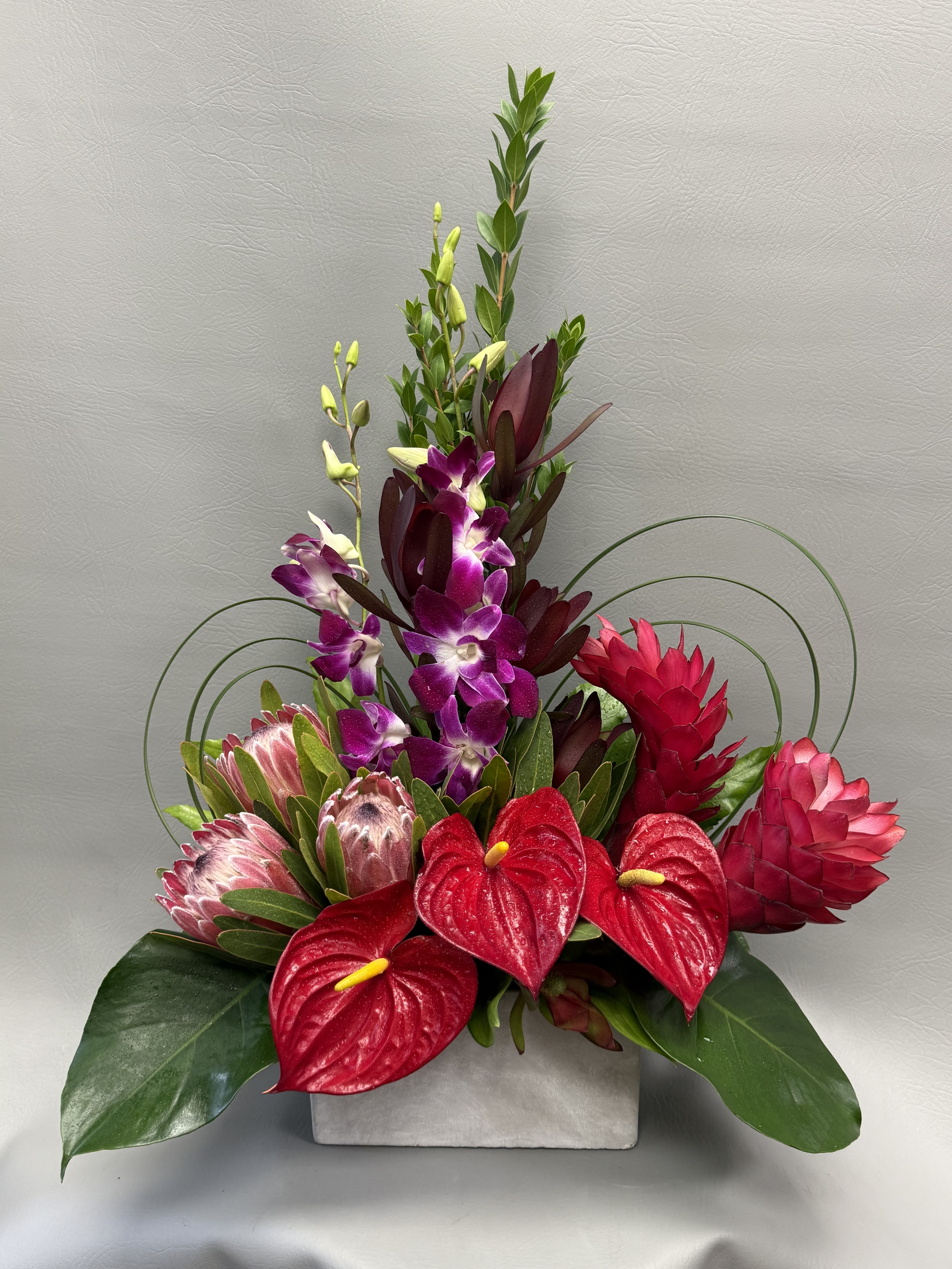 Tropical arrangement with red anthuriums, purple orchids, and pink protea in a white vase