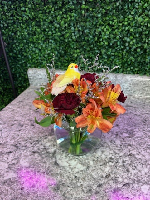 Bouquet of orange and burgundy flowers in a clear glass vase with a yellow bird figurine