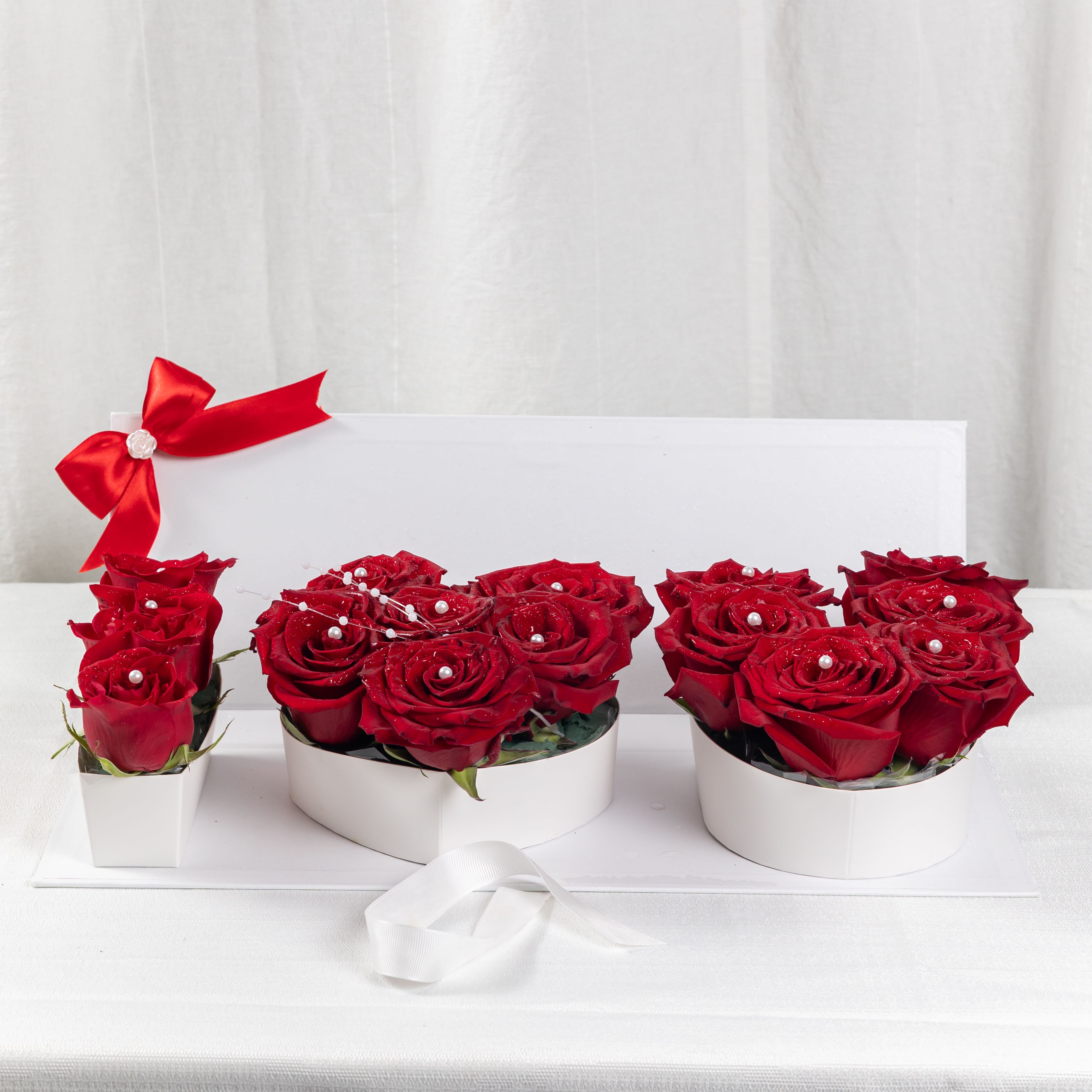 ETERNAL LOVE - A trio of red rose arrangements in heart, round, and rectangular white containers, adorned with delicate white beads and satin ribbons. A graceful display of love, perfect for intimate celebrations and timeless gestures.